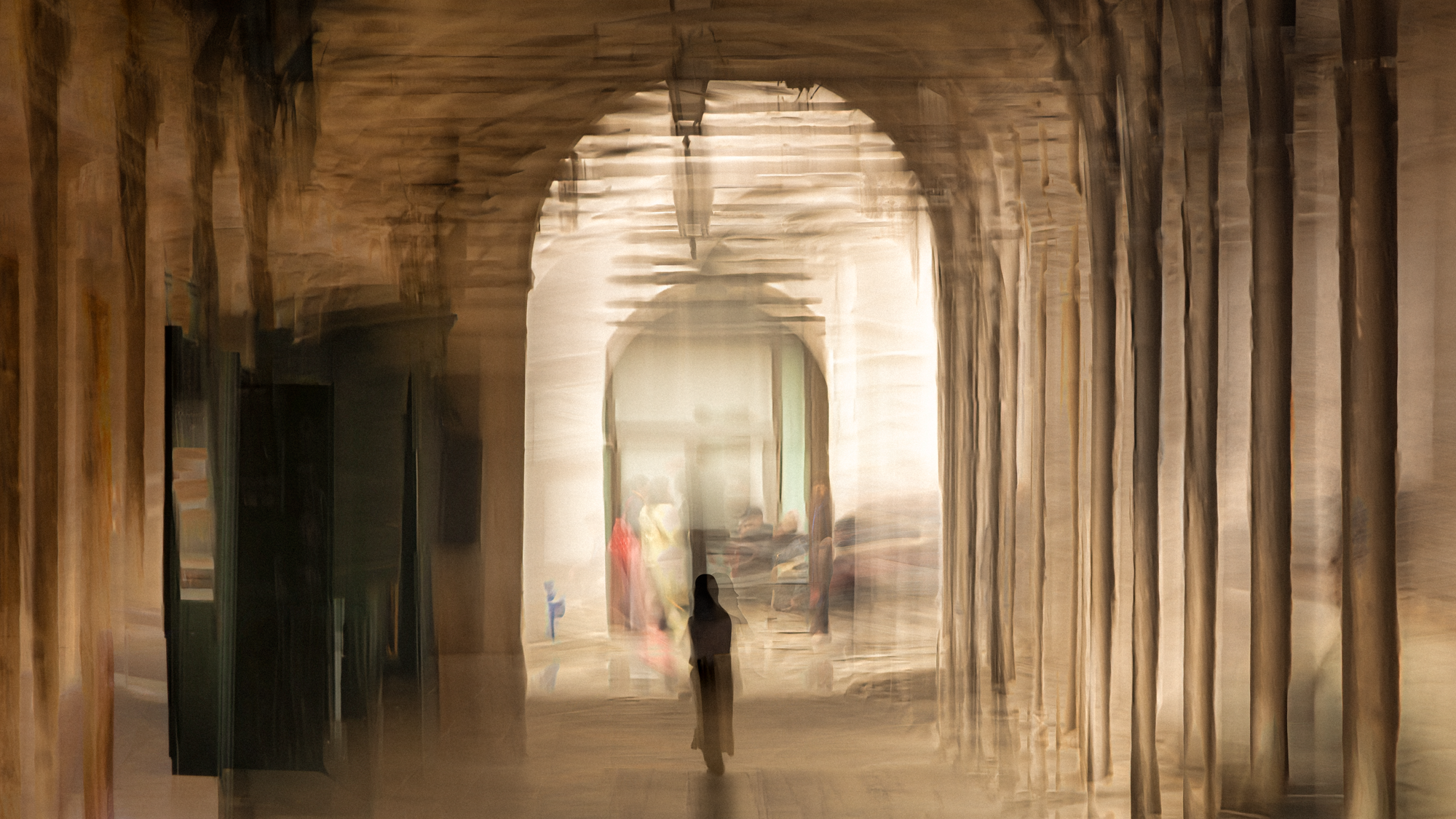 Impressionist ICM photograph of a solitary figure walking through a deep Venetian colonnade toward a bright archway, with blurred stone columns and distant hints of color in the light.