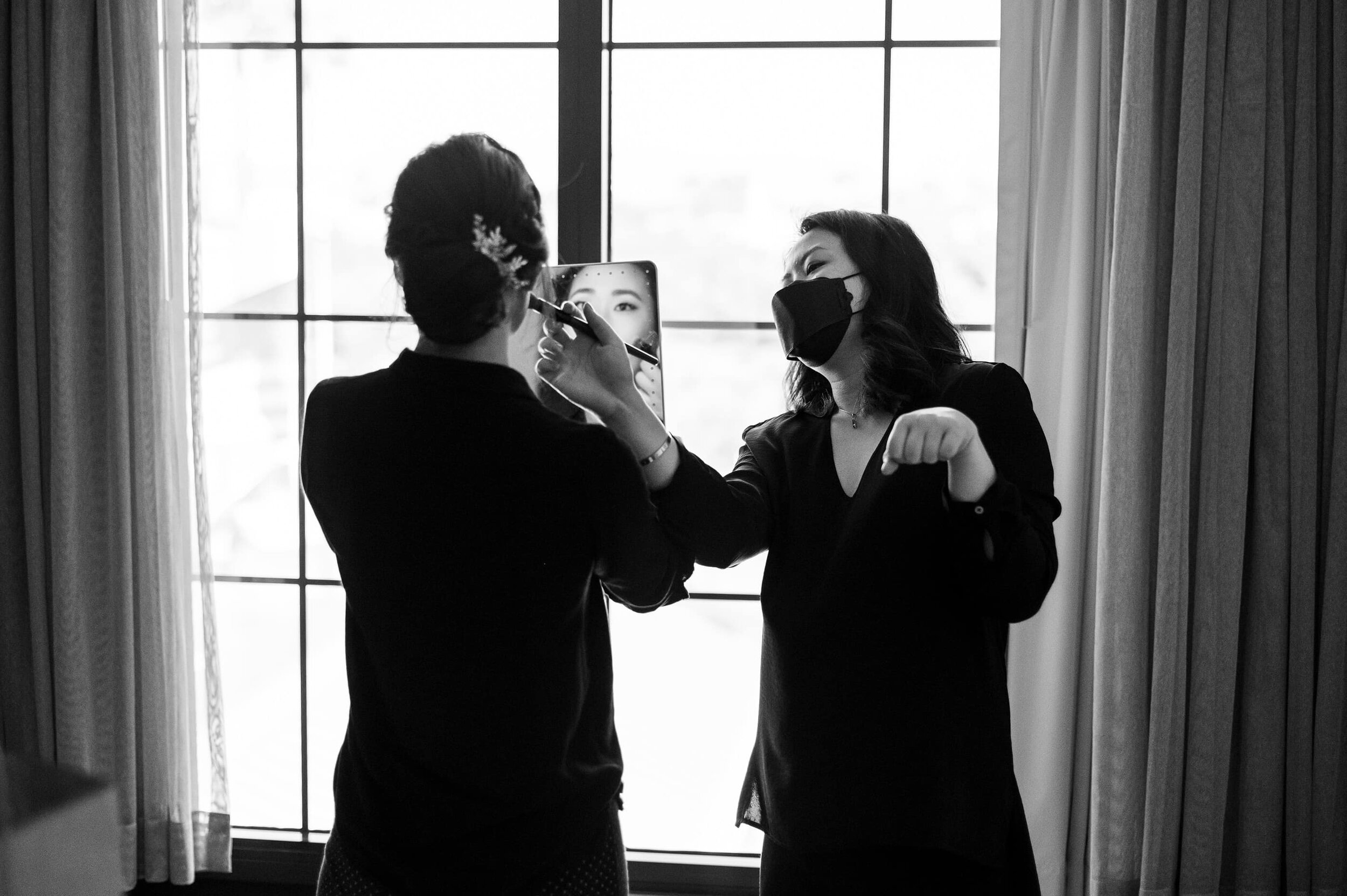 Black and white photo of bride getting make up applied while looking in mirror