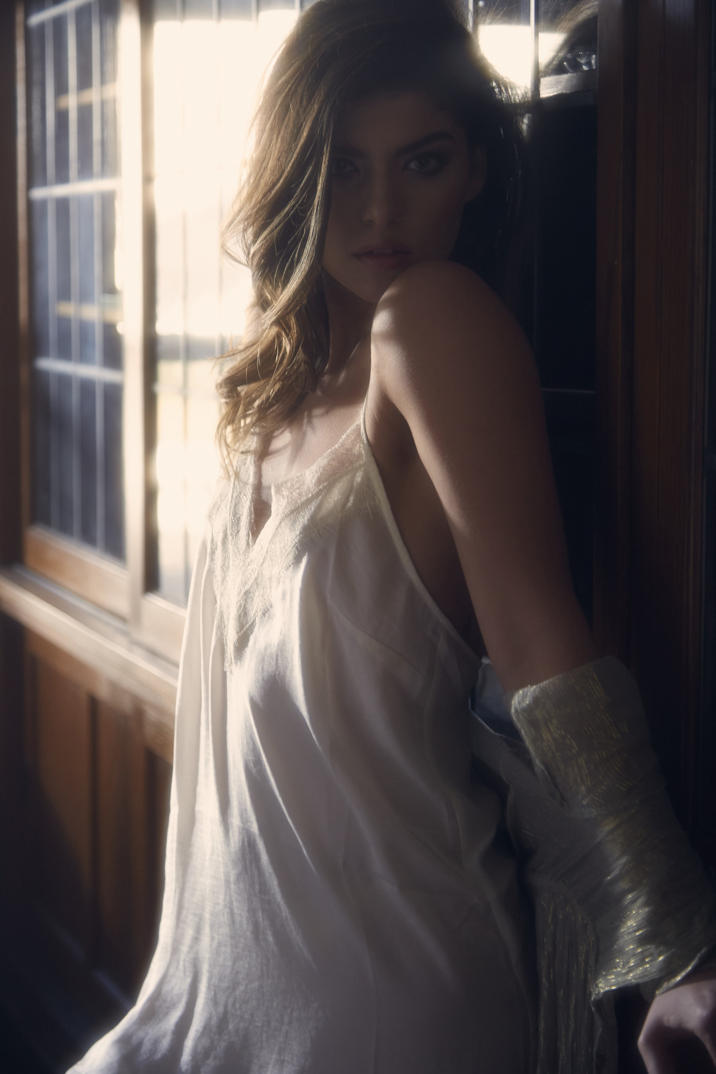  Romantic fashion editorial styled featuring model in white slip dress leaning against wall in moody natural light 