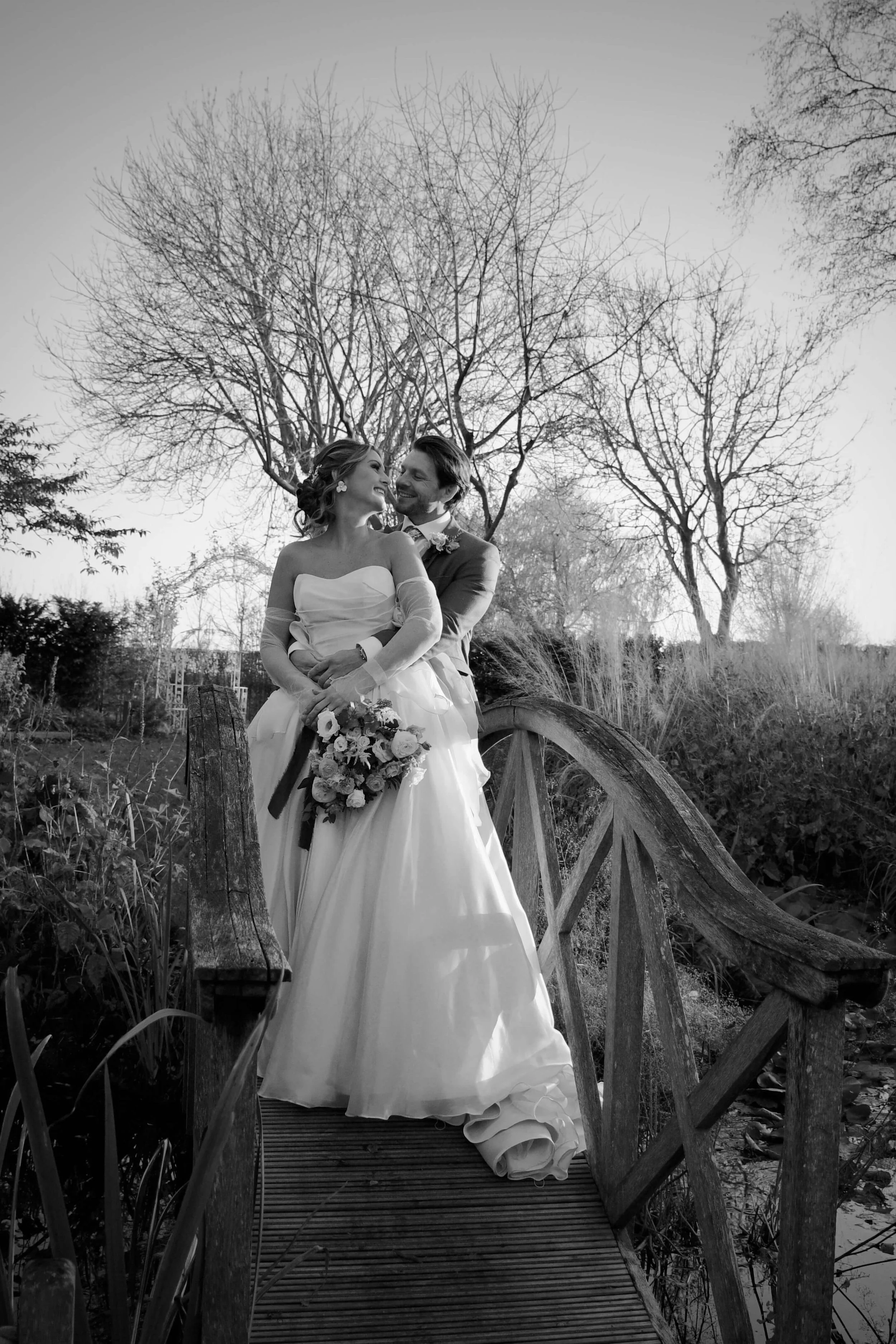 Candid wedding moment of a couple holding each other on a rustic bridge, captured in soft natural light in a black-and-white style.