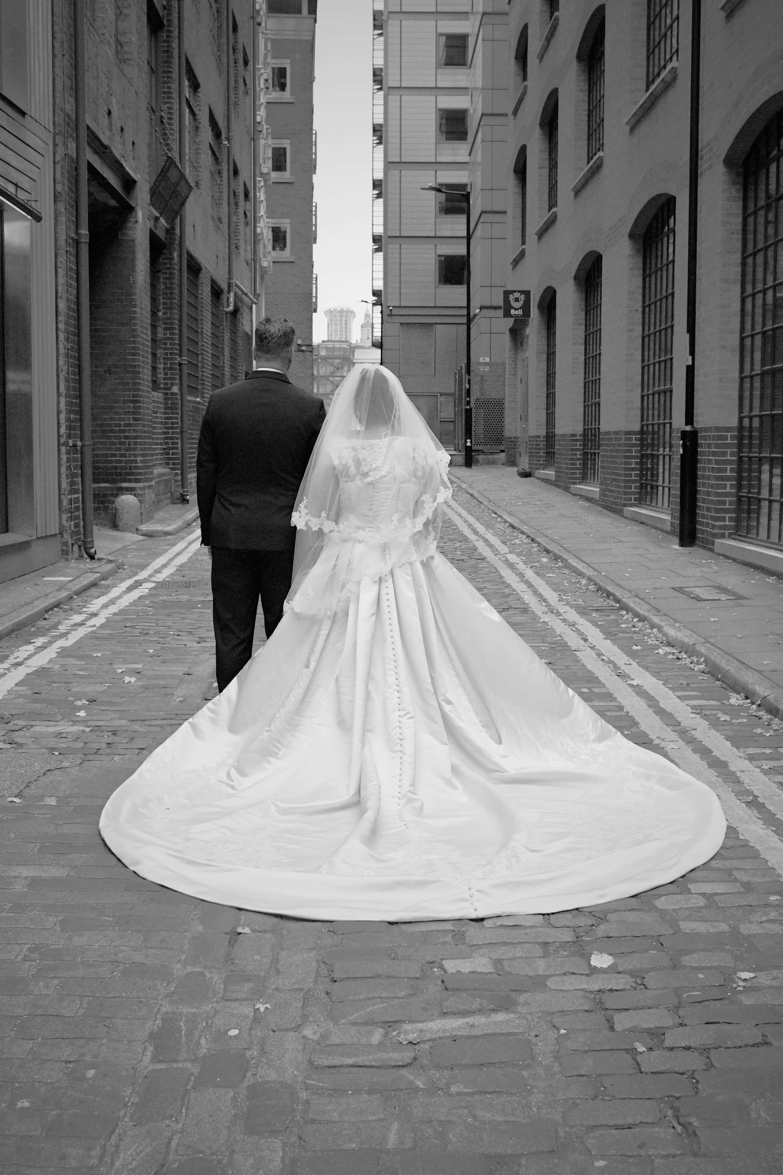 Documentary wedding photo of a couple walking down a cobblestone London alley, with the bride’s long train flowing behind her between historic brick buildings