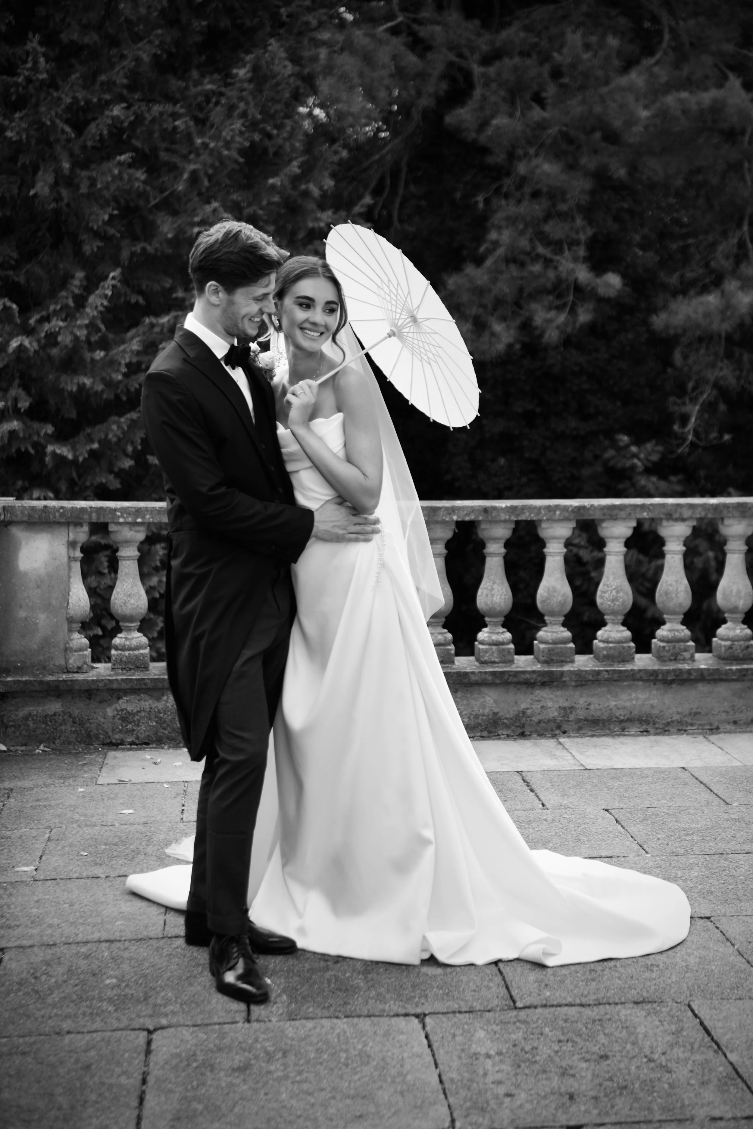 Black and white wedding portrait of bride and groom embracing beneath a white parasol on a garden terrace in London