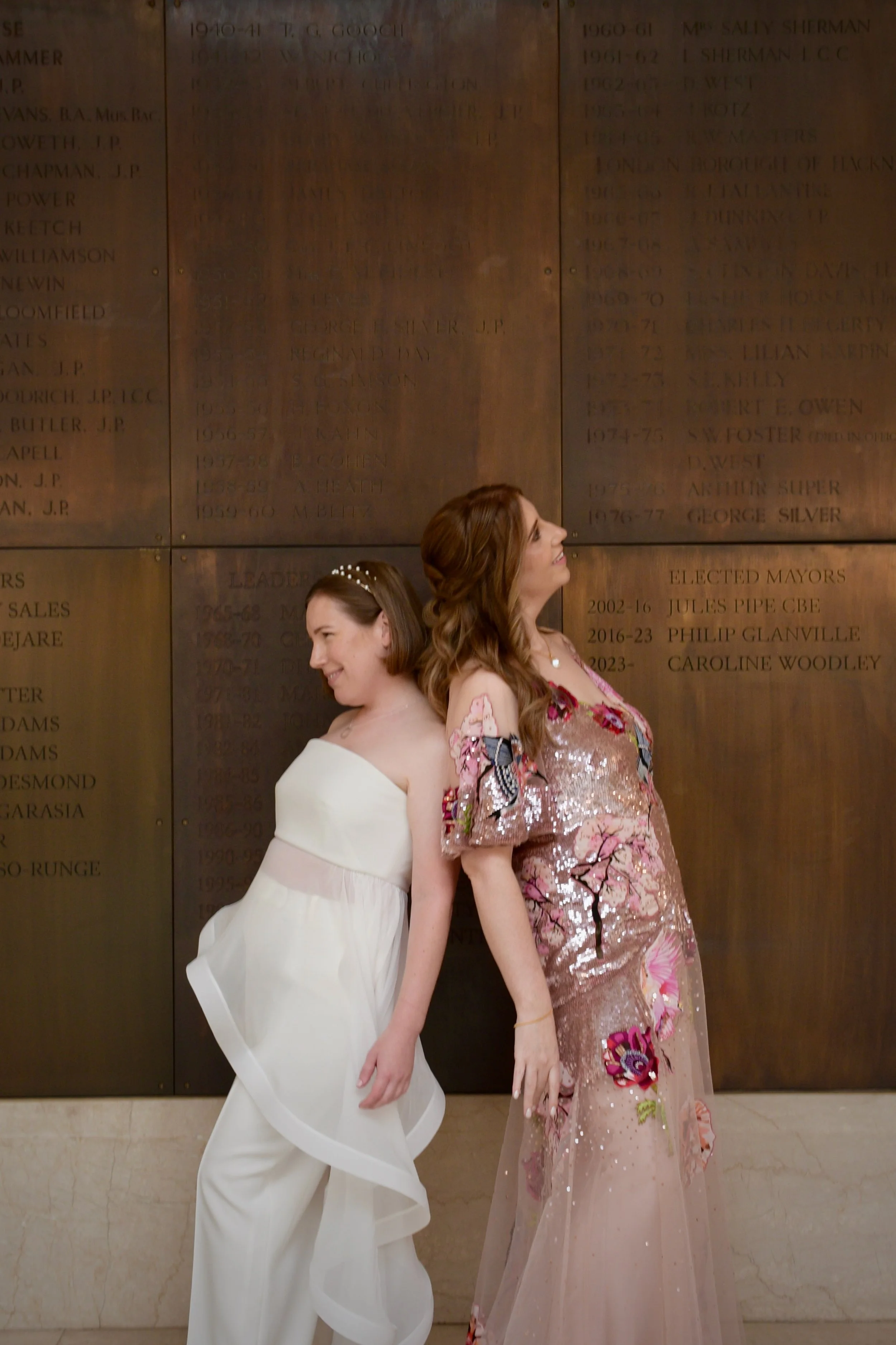 Same-sex wedding of two brides smiling back to back in elegant dresses at the historic Hackney Town Hall.