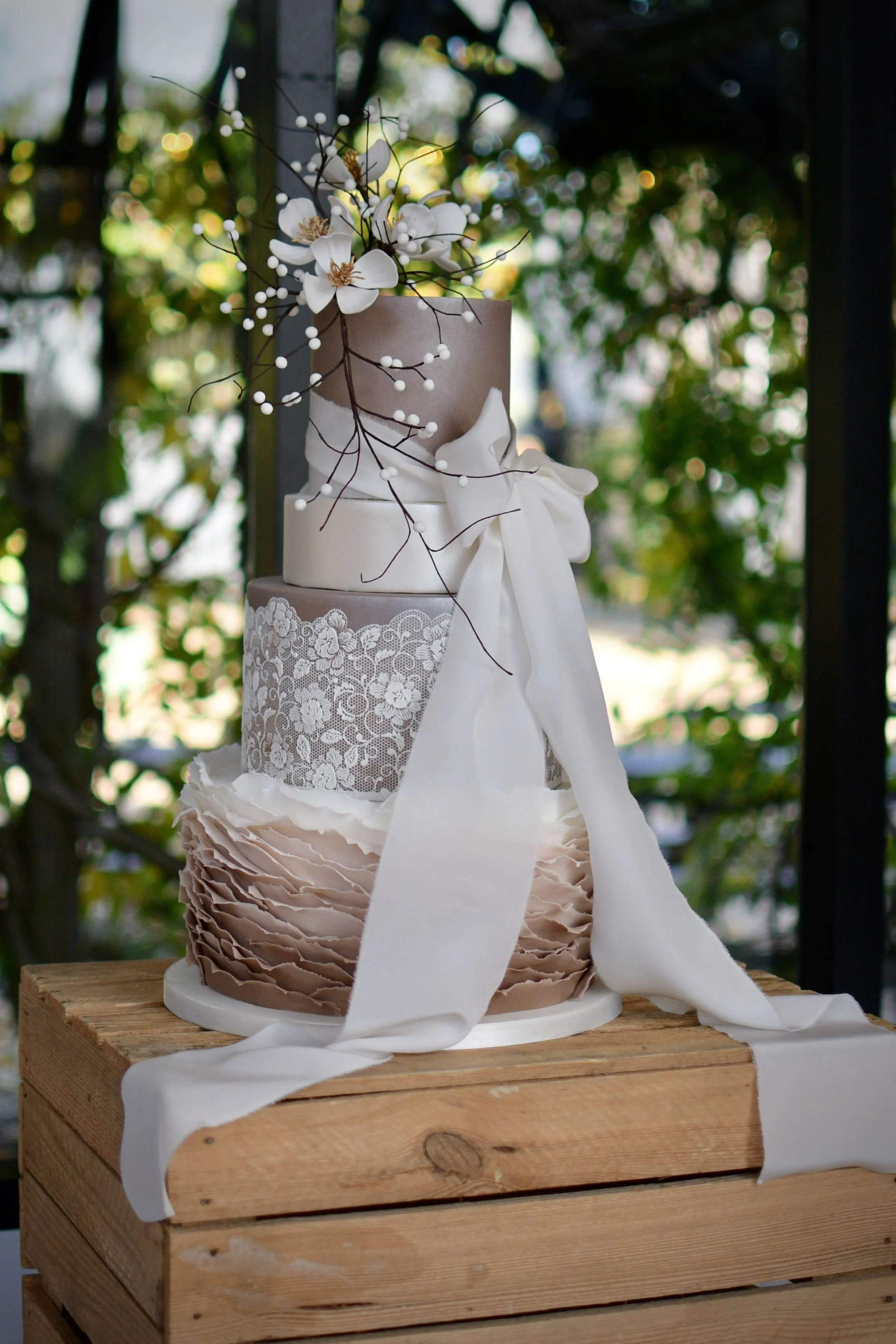Tall, multi-tiered wedding cake decorated with lace-inspired detailing, delicate sugar flowers, and flowing white ribbon, displayed on rustic wooden crates in natural outdoor light.