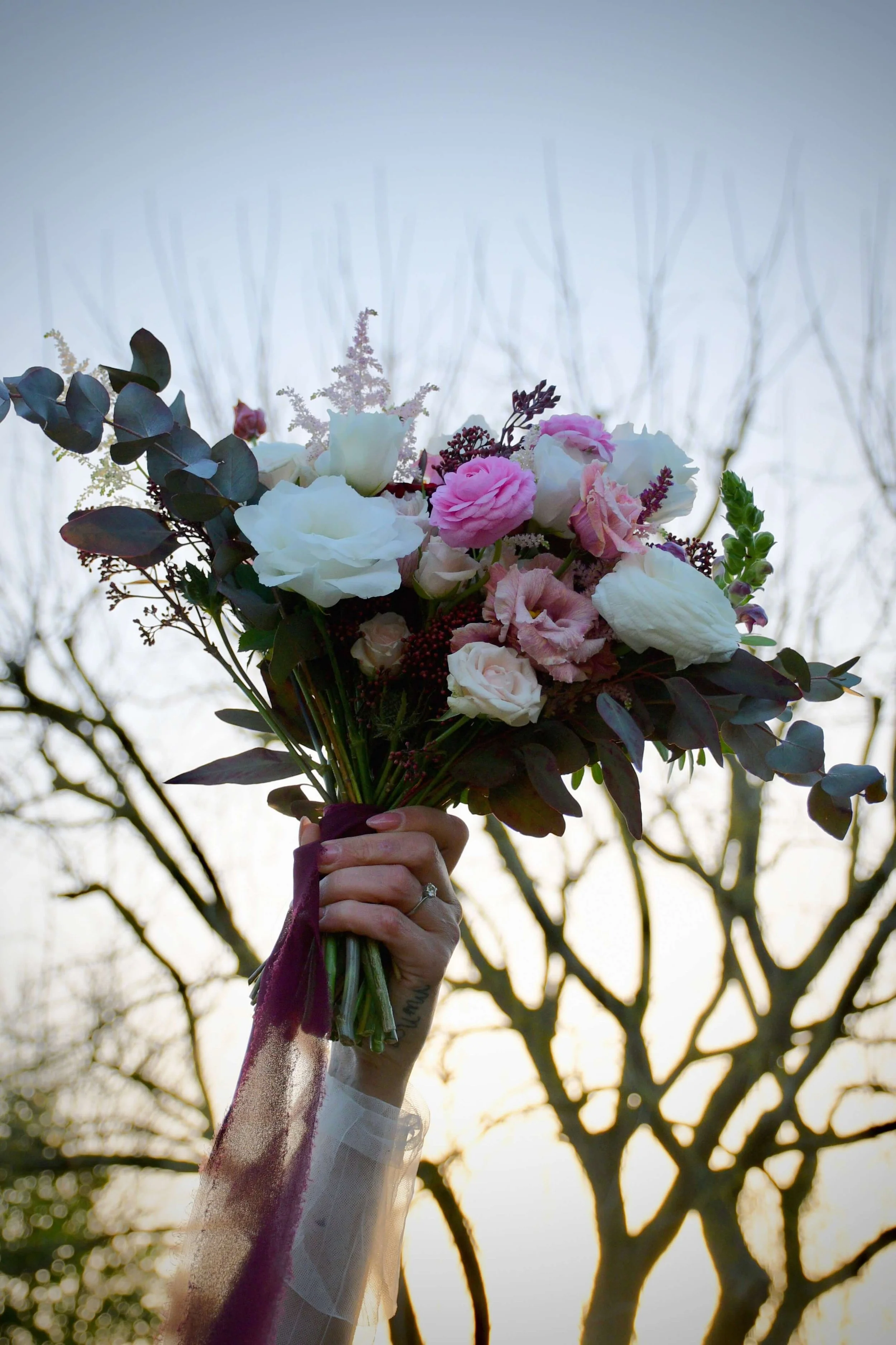Bride’s hand holding a romantic bouquet of pink and white roses, ranunculus, and eucalyptus, photographed at sunset with bare winter trees in the background.