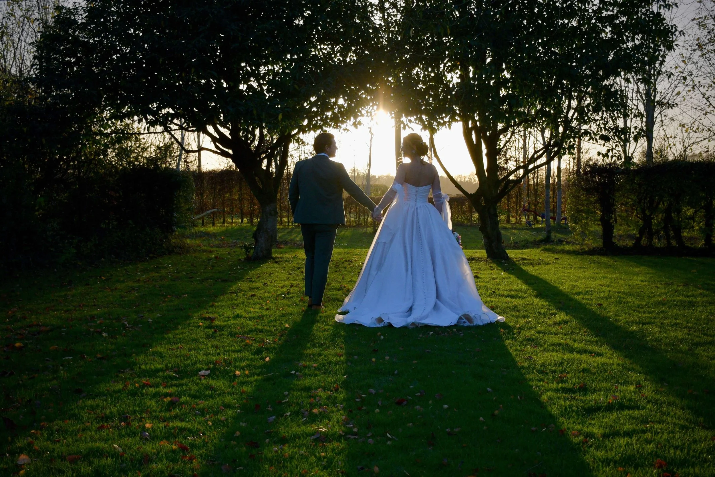 Documentary wedding photo of a couple walking hand in hand across a sunlit garden at golden hour, with long shadows and natural greenery surrounding them.
