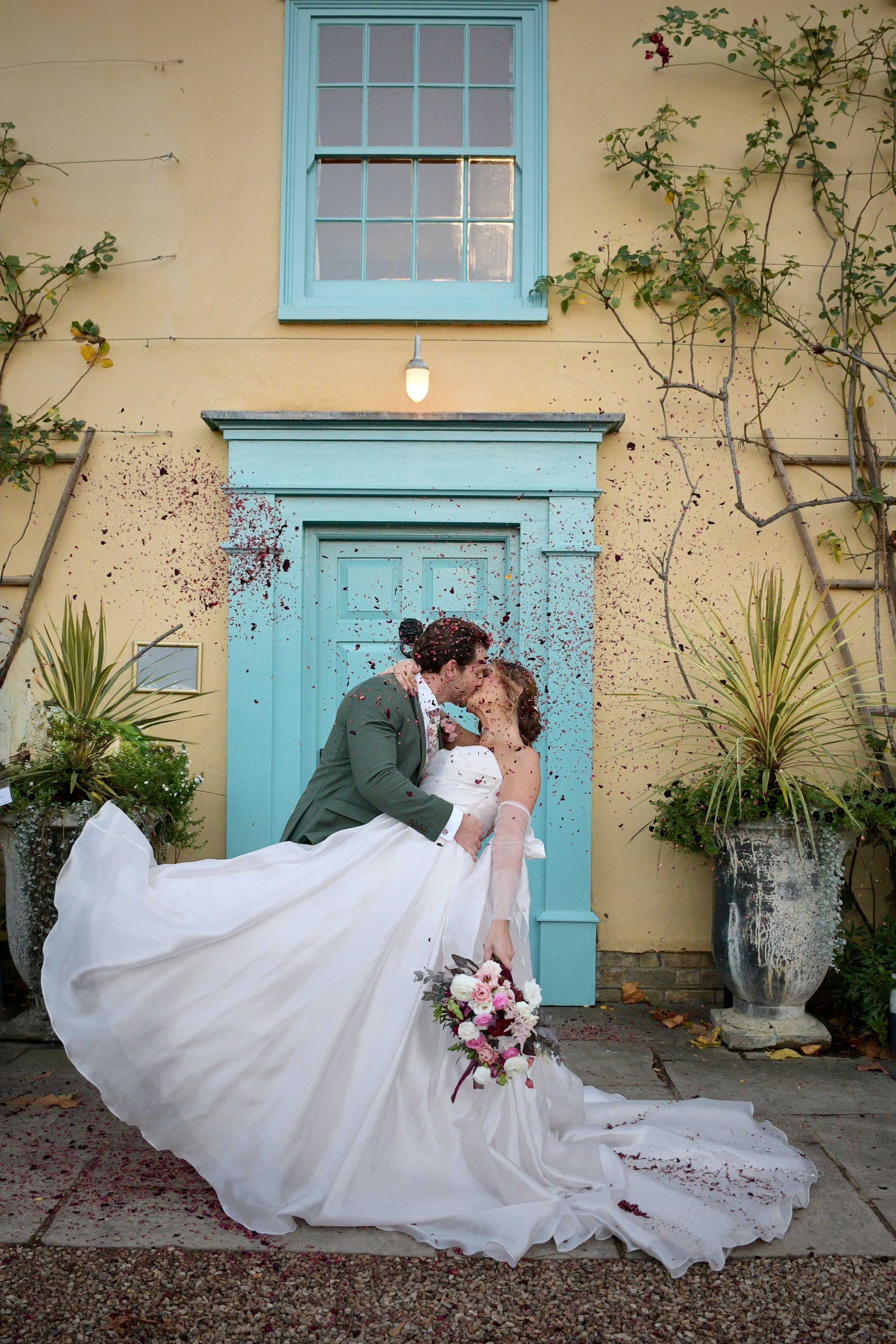 Bride and groom kissing under falling confetti in front of a pastel blue doorway during a romantic outdoor wedding.
