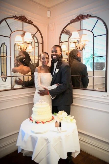 Bride and groom posing beside their wedding cake in an elegant London wedding venue with decorative mirrors reflecting the couple.