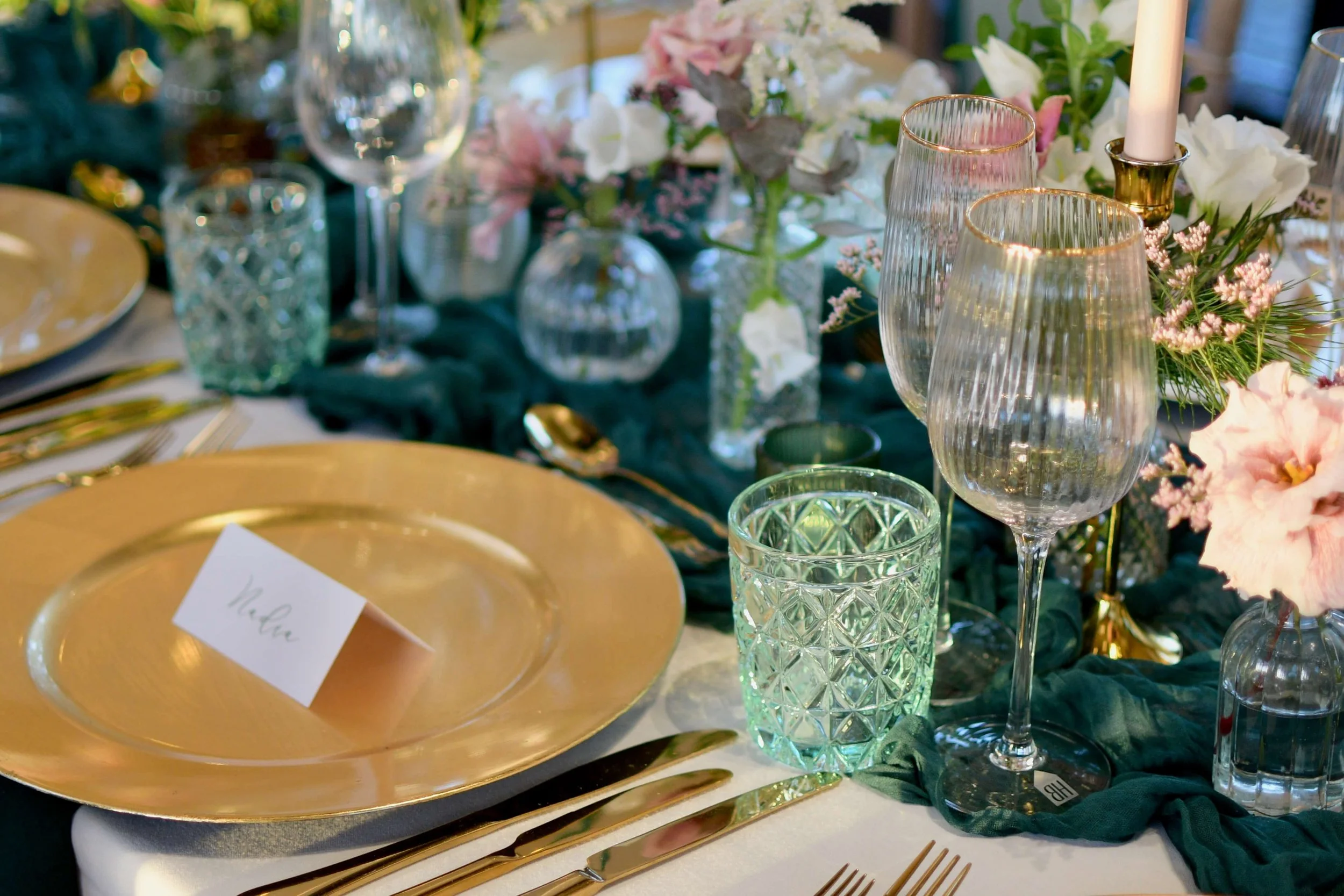 Elegant wedding table setting featuring gold charger plates, crystal wine glasses, pastel floral arrangements, and luxurious green fabric décor, captured in soft natural light.