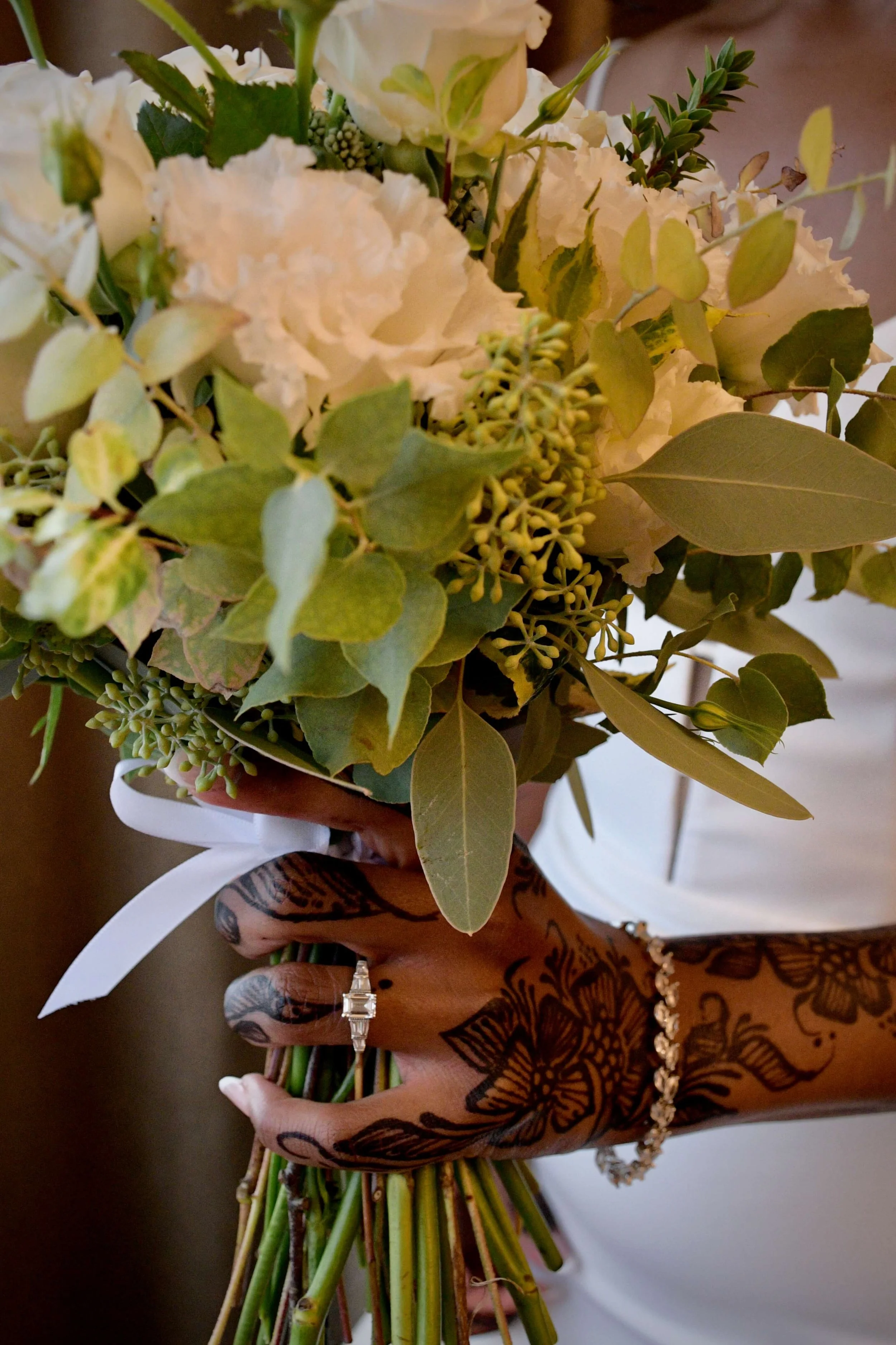 Close-up of a bride's hand adorned with intricate dark brown henna tattoos, holding a lush white and green wedding bouquet with a white ribbon. A sparkling diamond engagement ring and bracelet are visible, highlighting traditional bridal beauty and e