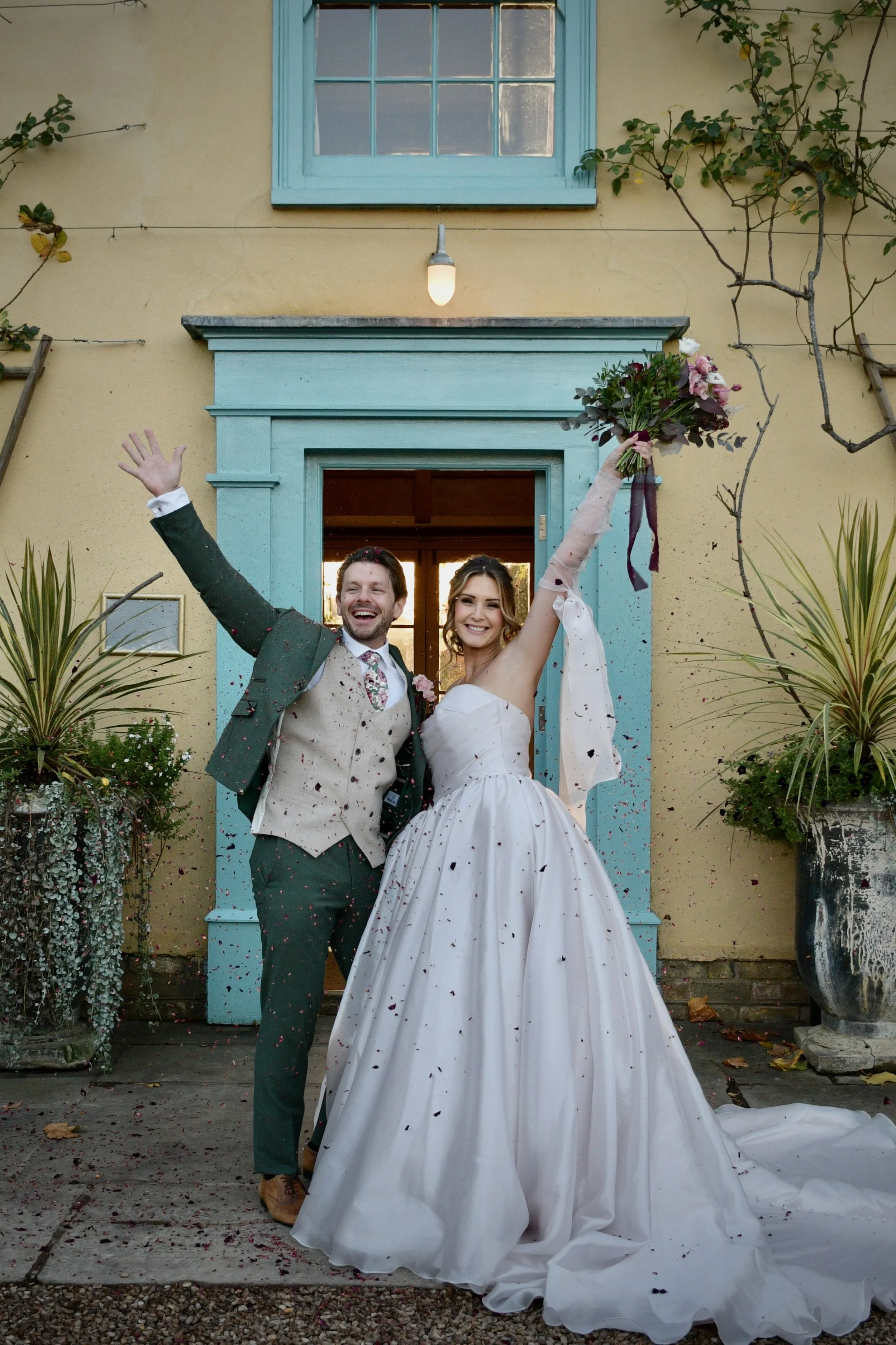 Bride and groom celebrating outside venue with bouquet raised and confetti falling.