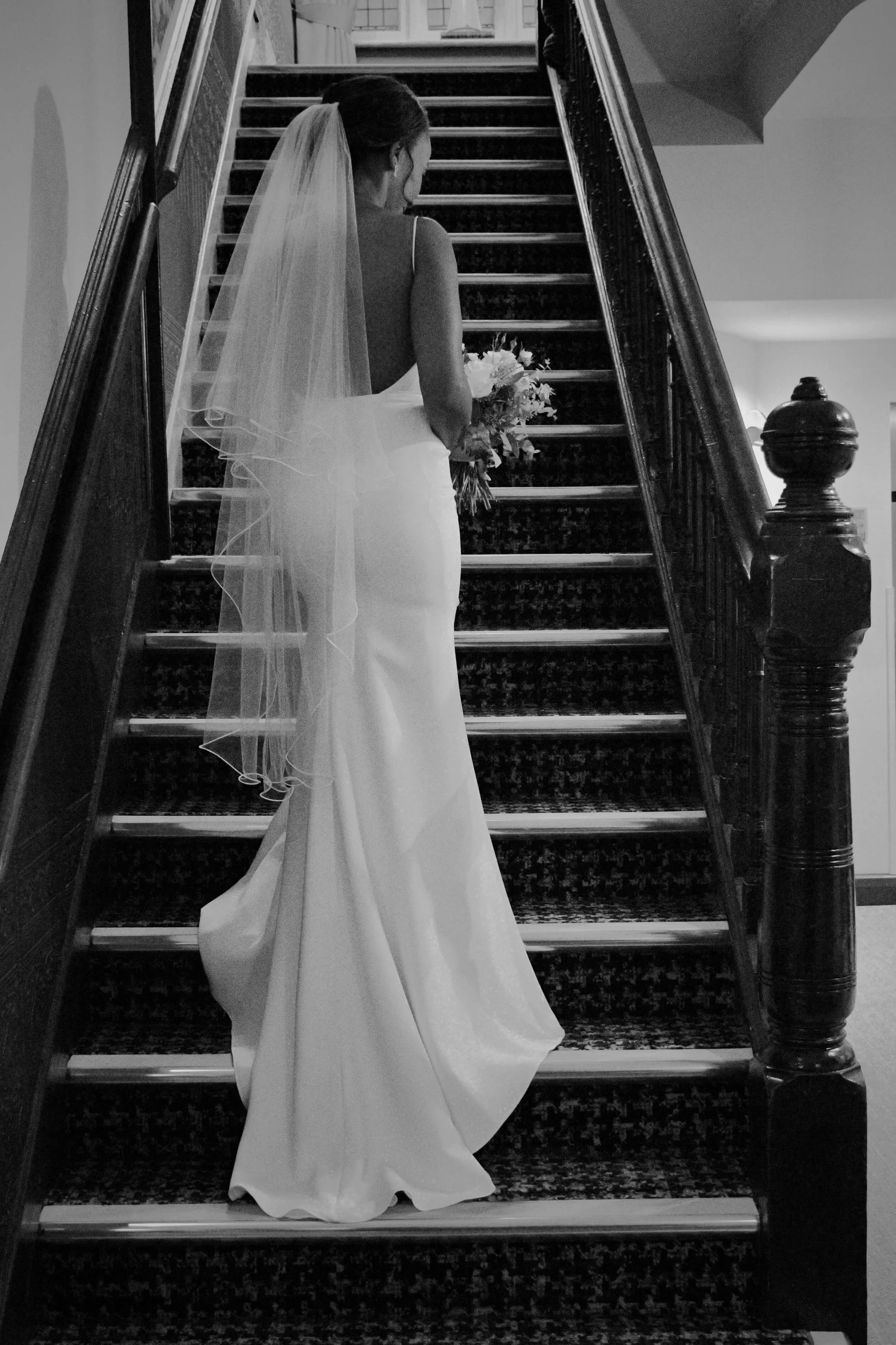 Timeless black and white bridal portrait of a bride ascending a staircase in a modern minimalist wedding gown and veil, holding a bouquet at an elegant indoor London wedding venue.