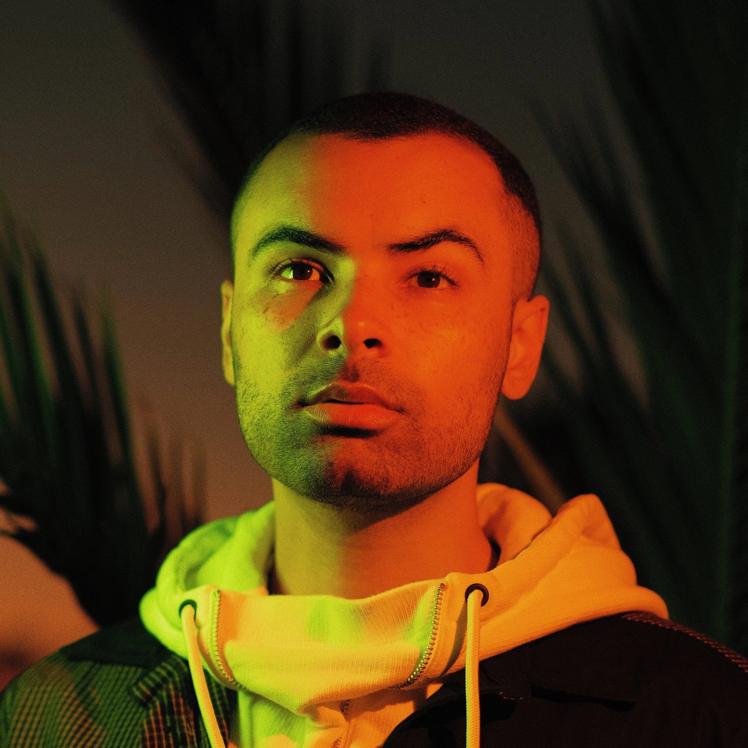 EHN JEY, illuminated with multicolored lighting, wearing a white hoodie and a dark jacket, with a background of dark, leafy plants.