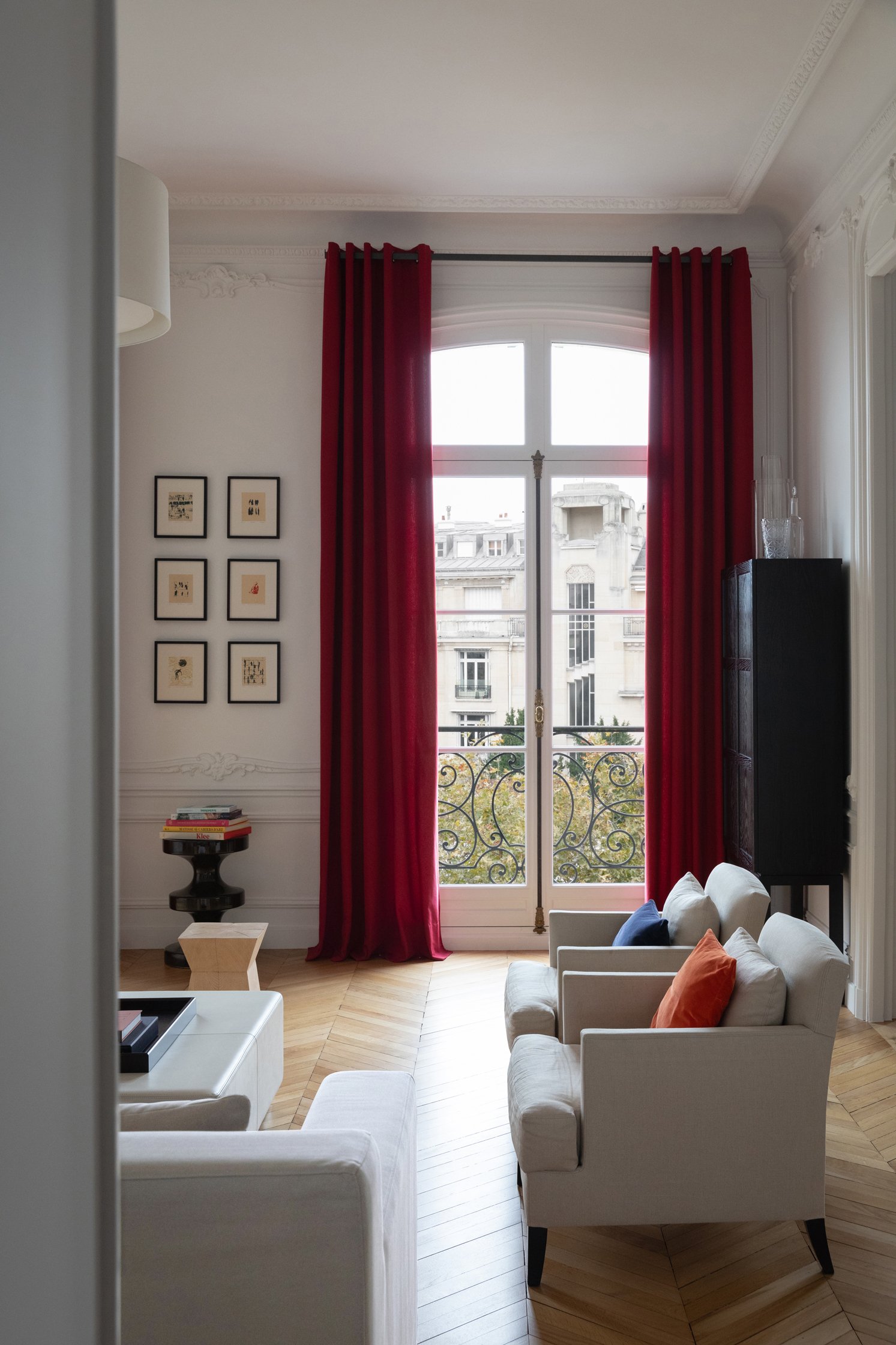 Salon d’un appartement haussmannien rénové avec grandes fenêtres, rideaux rouges et parquet en point de Hongrie