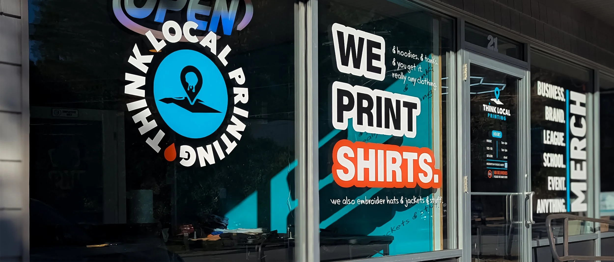 Store window advertising local printing shirts, hoodies, tanks, embroidered hats and jackets, and other items, with text 'We Print Shirts.' and a 'Think Local Printing' sign.