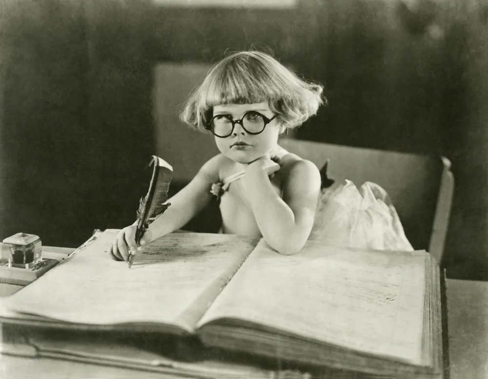 Young girl with glasses writing with a quill in a large book