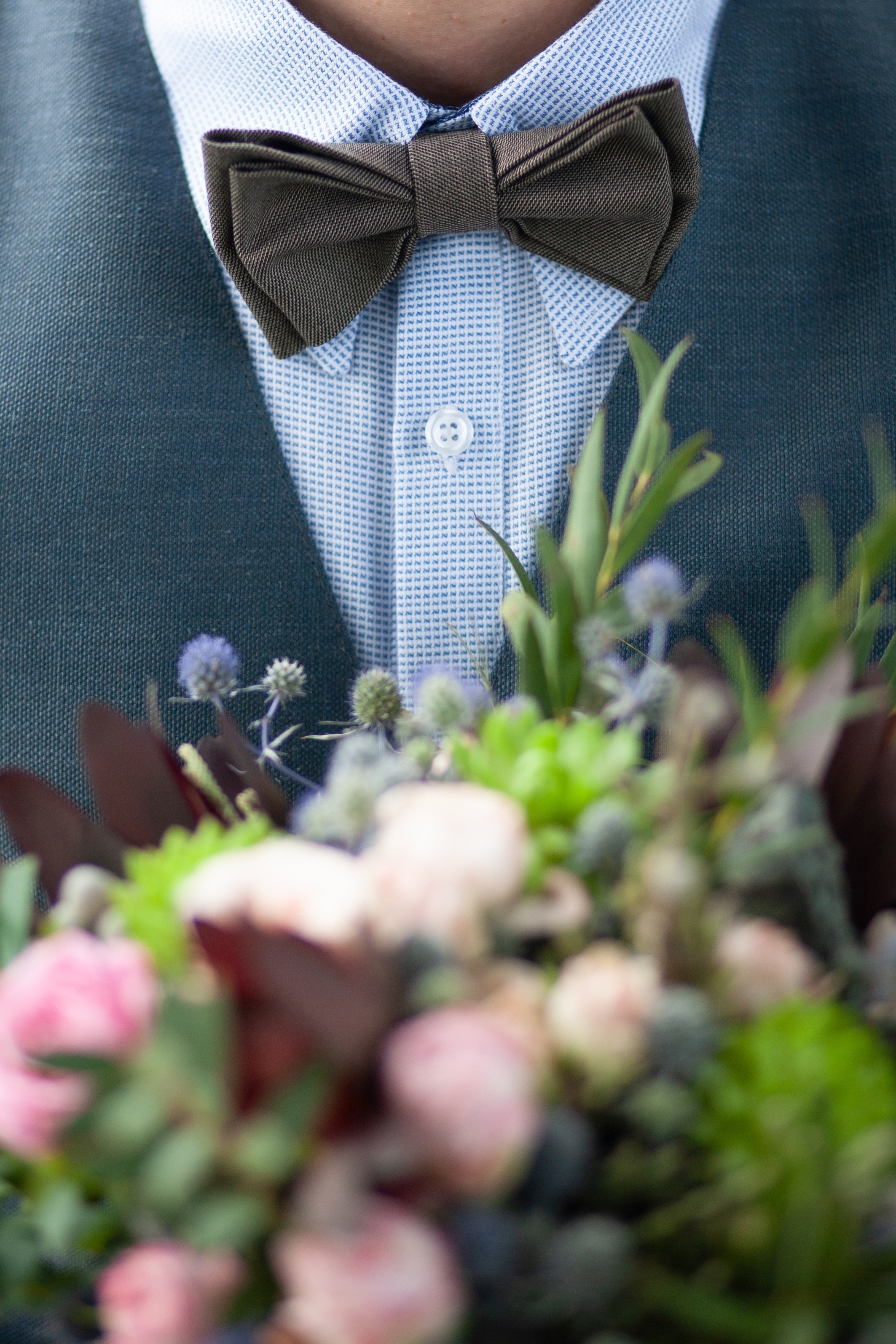 Beautiful rustic style in wedding decor. Closeup view of groom wearing blue shirt, vest and brown bowtie holding wedding flowers in hands. Vertical color photography.