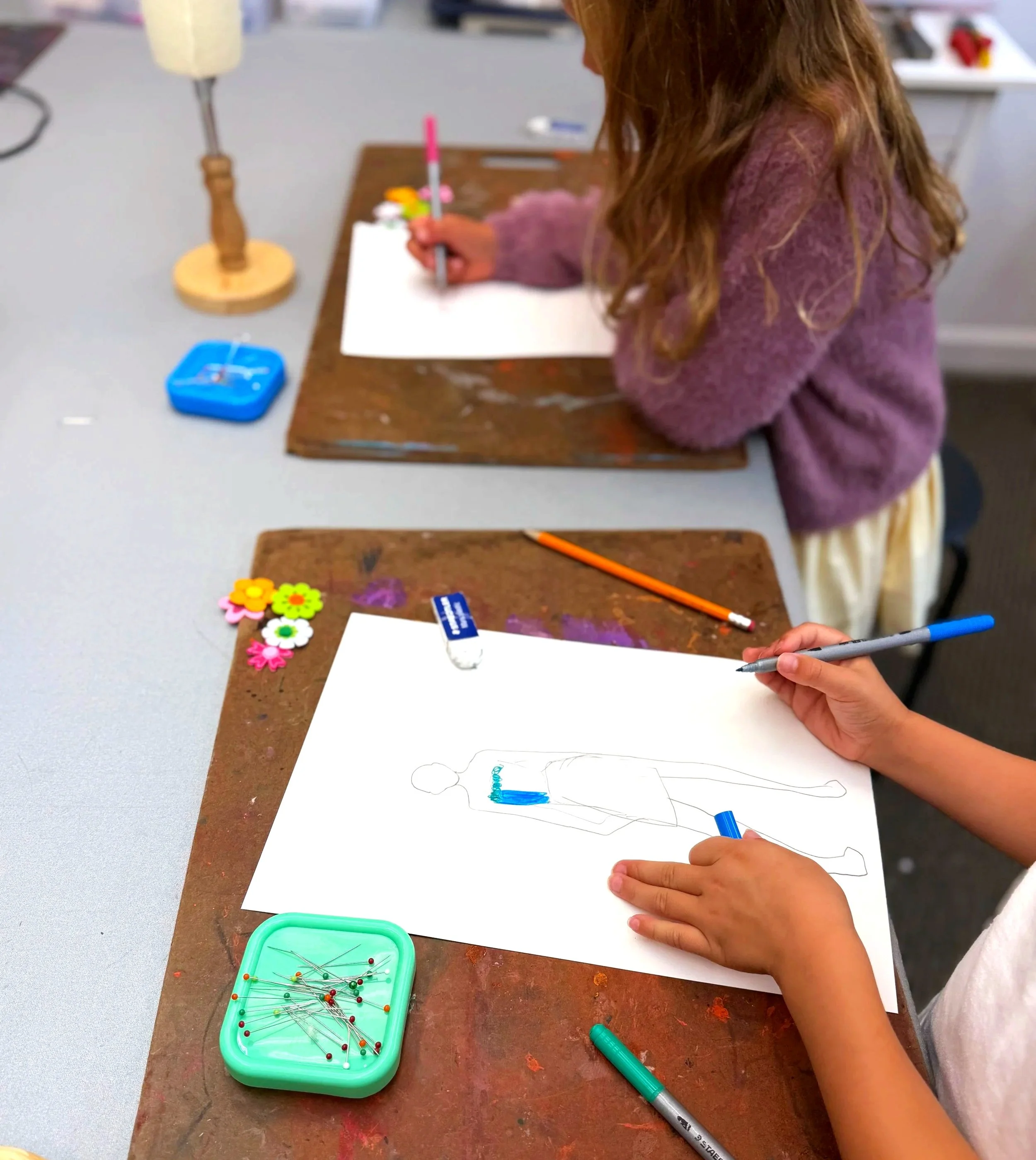 Girls in lavender sweater and long hair drawing with colored pencils and markers on a drawing board on a table.
