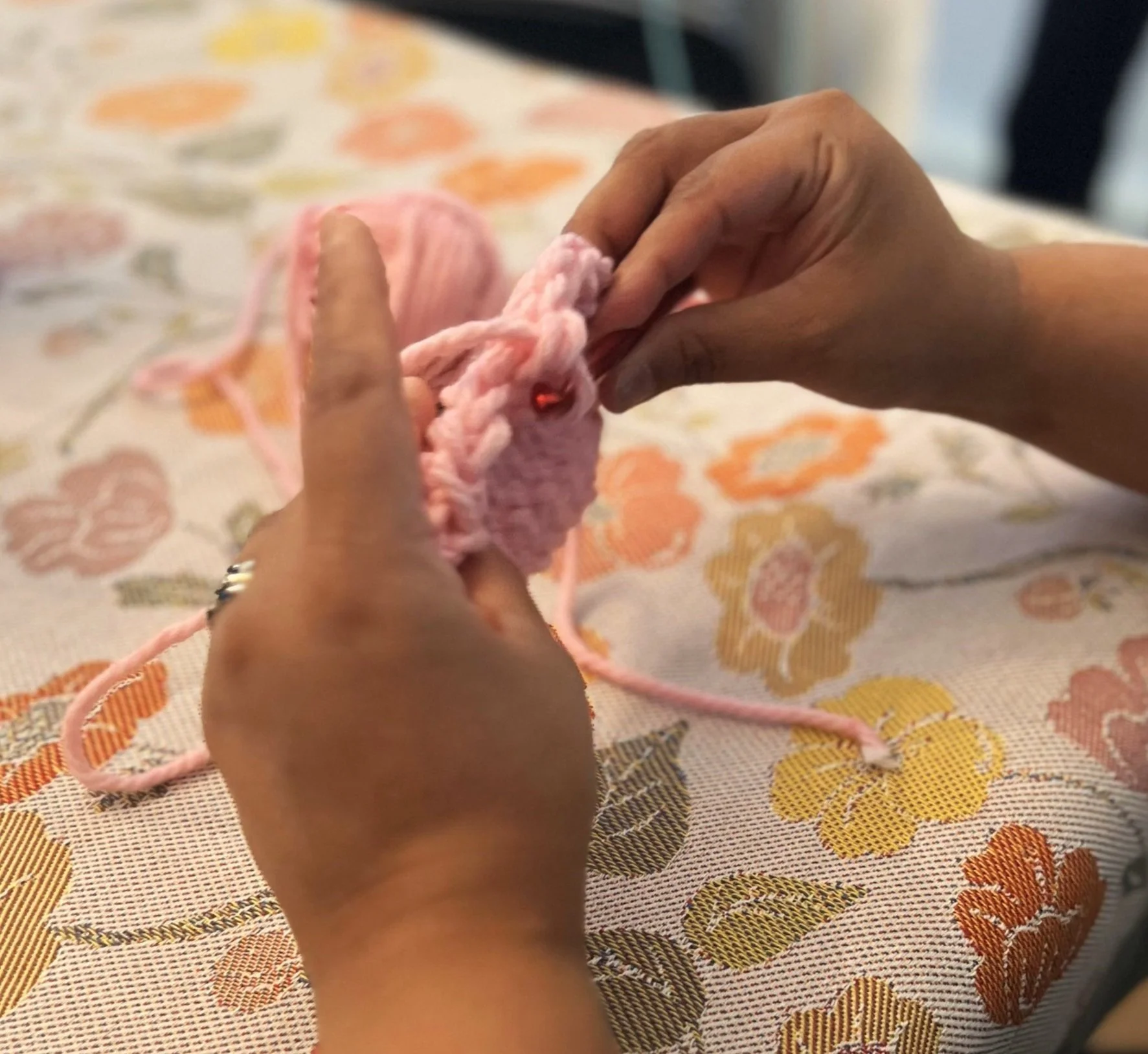 Hands knitting with pink yarn on a floral tablecloth.