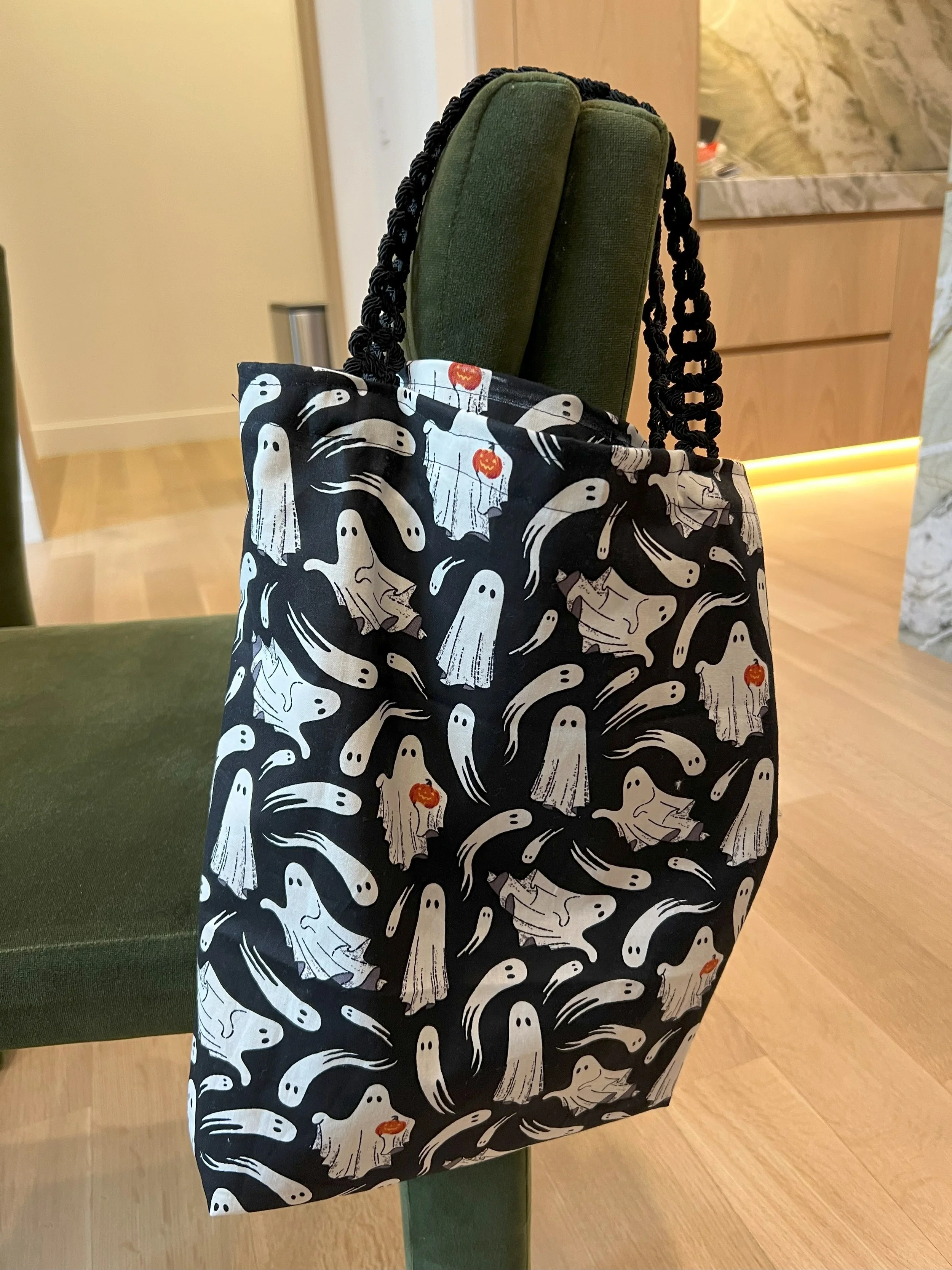 Halloween-themed tote bag hanging on a green chair with a design of white ghosts and small orange pumpkins on a black background.