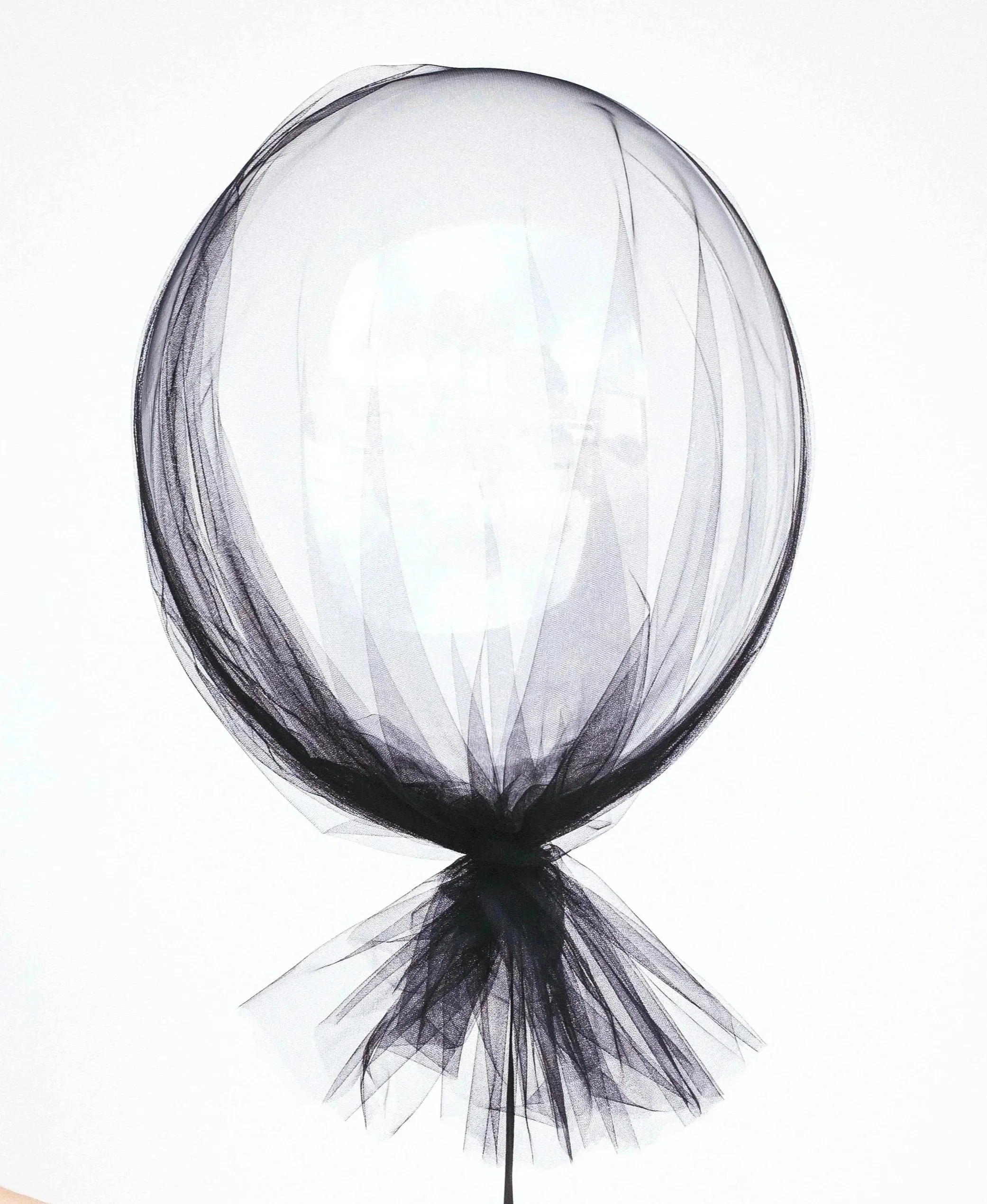 A black and white image of a balloon made of layered transparent material, tied at the bottom with a small knot and reflected on a surface.