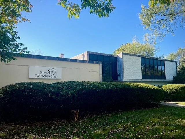 White Mid century modern building in New Providence, NJ, with large windows and green bushes in front, with a white sign that says Sew Dandelions