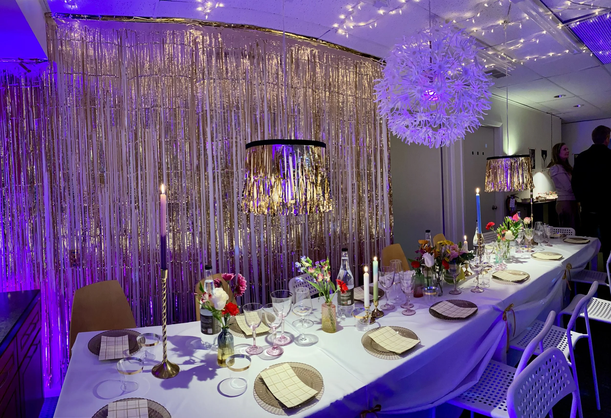 Long white clothed table set for party with glasses and plates, candles, and a wall covered with gold sparkle streamers.
