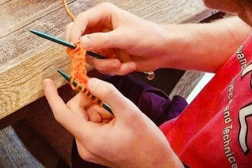 man's hands knitting with green needles and orange yarn