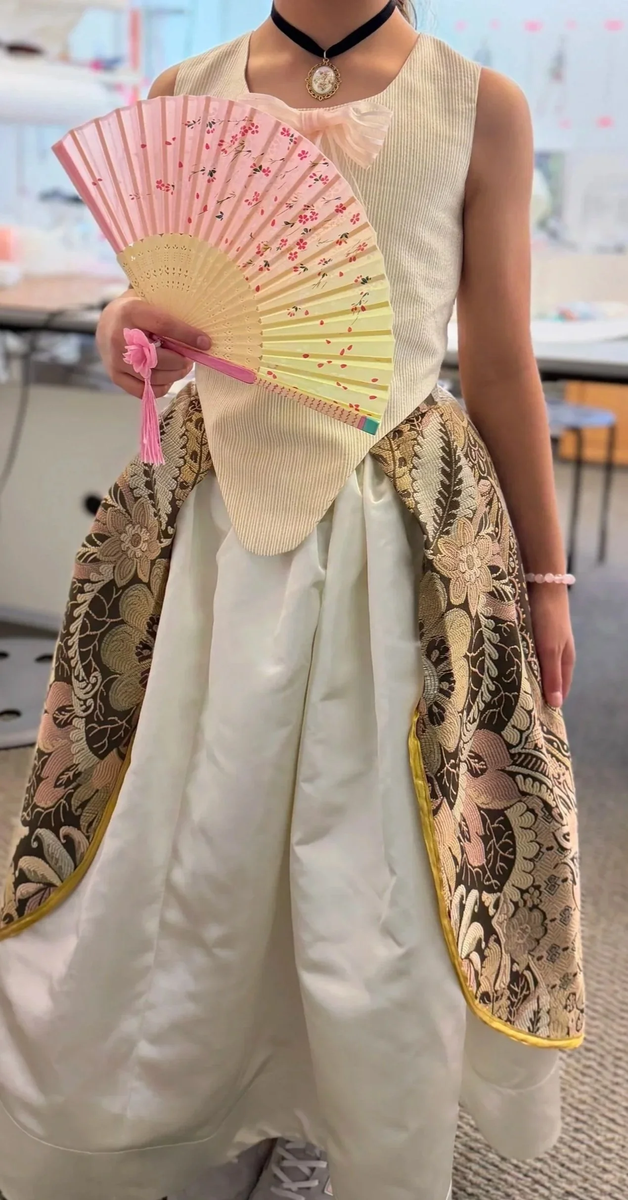 Young girl standing in Rococo costume of white gown, flower side skirt, choker, and a pink and yellow fan