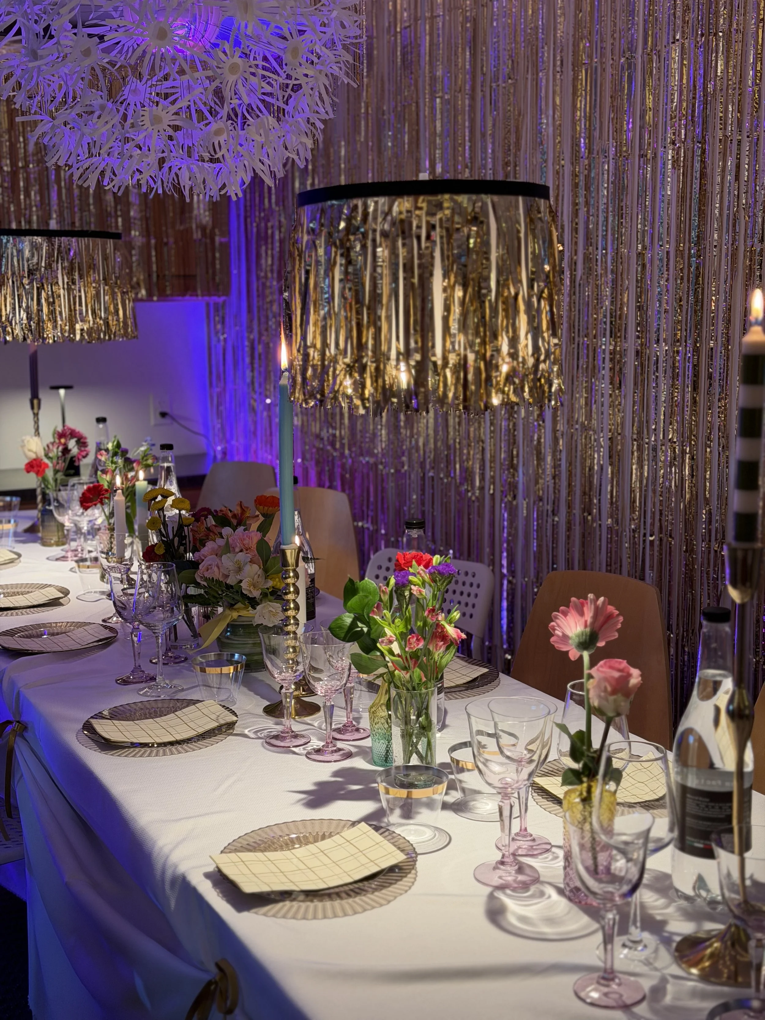 Long white clothed table set for party with glasses and plates, candles, and a wall covered with gold sparkle streamers. Multiple vases with fresh flowers.