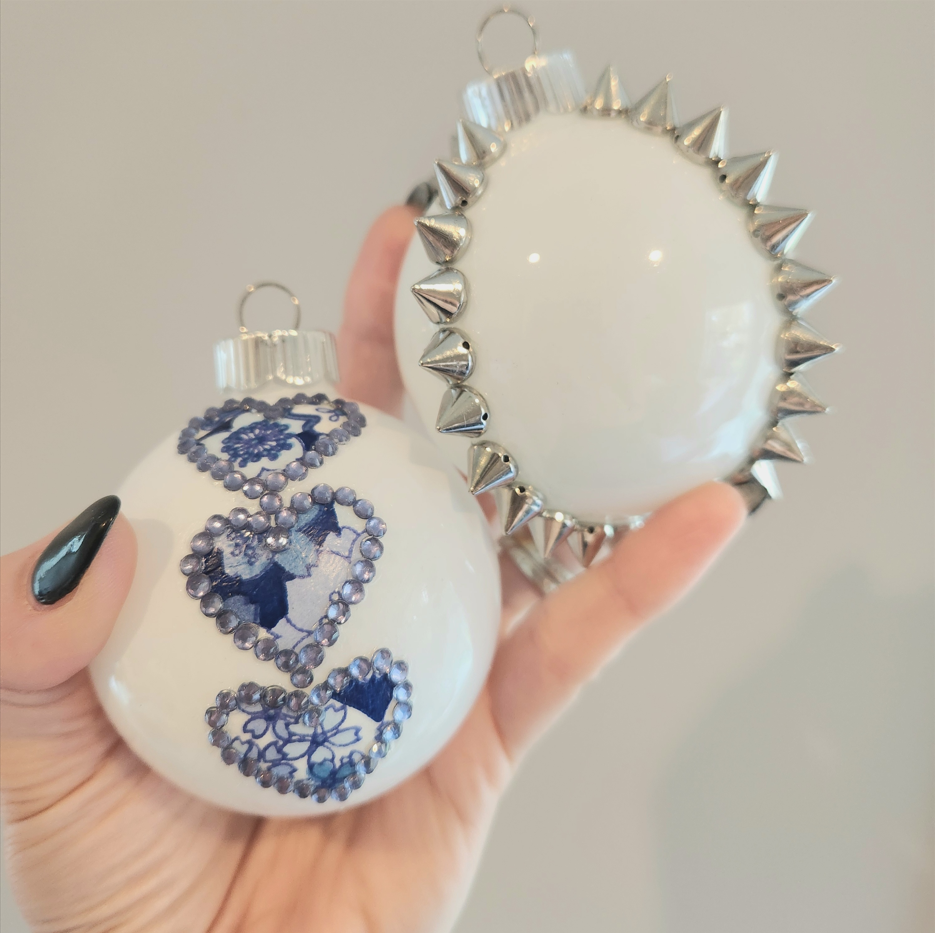 Two white Christmas ornaments, one decorated with blue sequins spelling 'Love' and the other with silver spikes around its perimeter, being held in a person's hand.