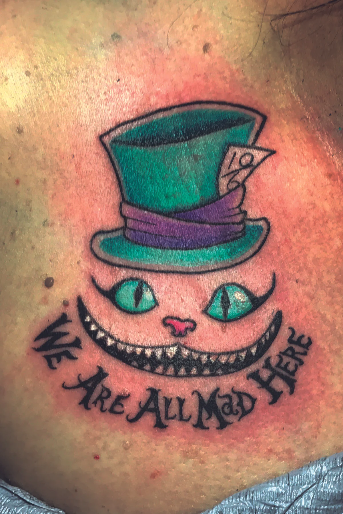 Cheshire cat smile and top hat, 'We are all mad here' in color. Below the collarbone tattoo. 