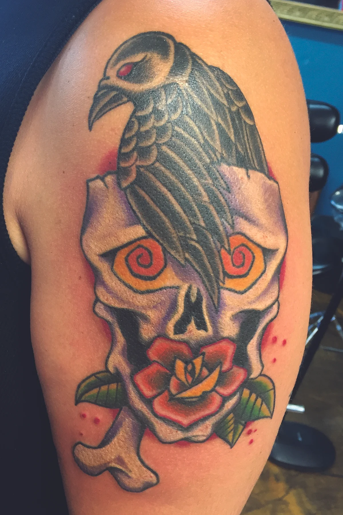 Black crow out of a skull with a rose and bone in color. Upper shoulder half arm sleeve tradational tattoo.