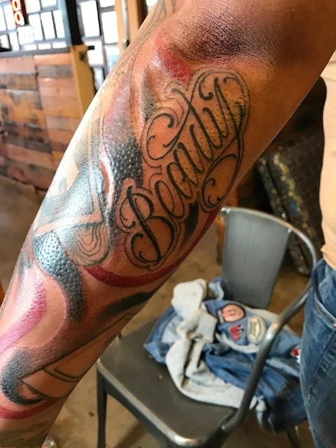 "Beauty" beautifully written in black and gray with a swirl of red white and blue on forearm. Full sleeve tattoo