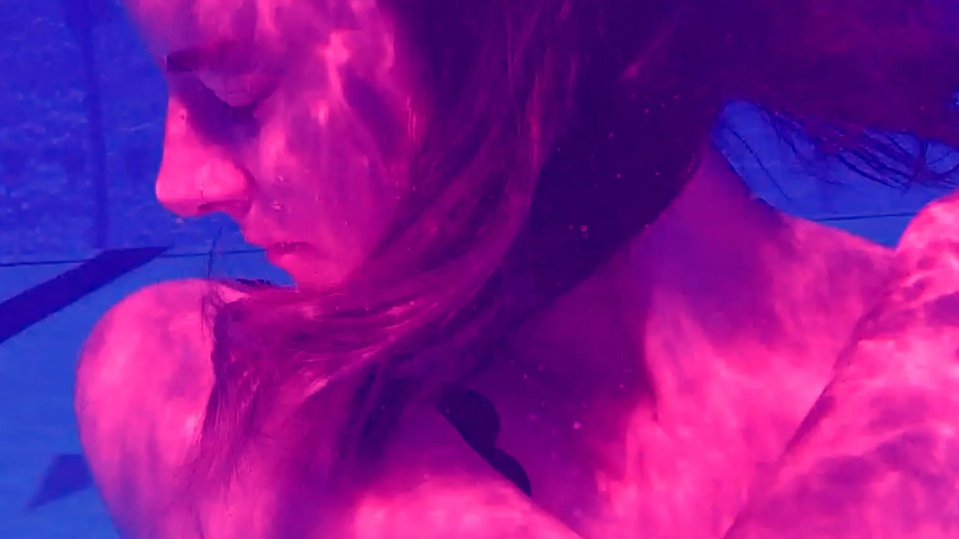 Close-up of a person underwater, shown in profile from the shoulders up. Long hair surrounds their face. Small air bubbles cling to their skin. The image is heavily tinted in vivid magenta and purple tones, contrasting against the bright blue underw