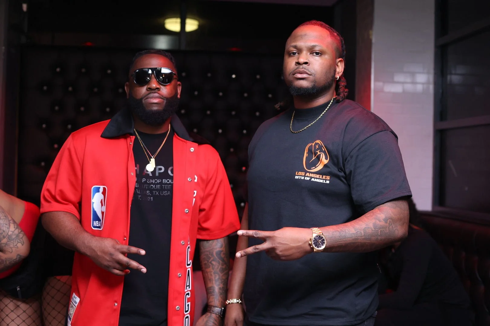 Two men standing indoors, both wearing black shirts, with the man on the left in a red jacket and sunglasses, and the man on the right with tattoos on his arm and showing a peace sign.