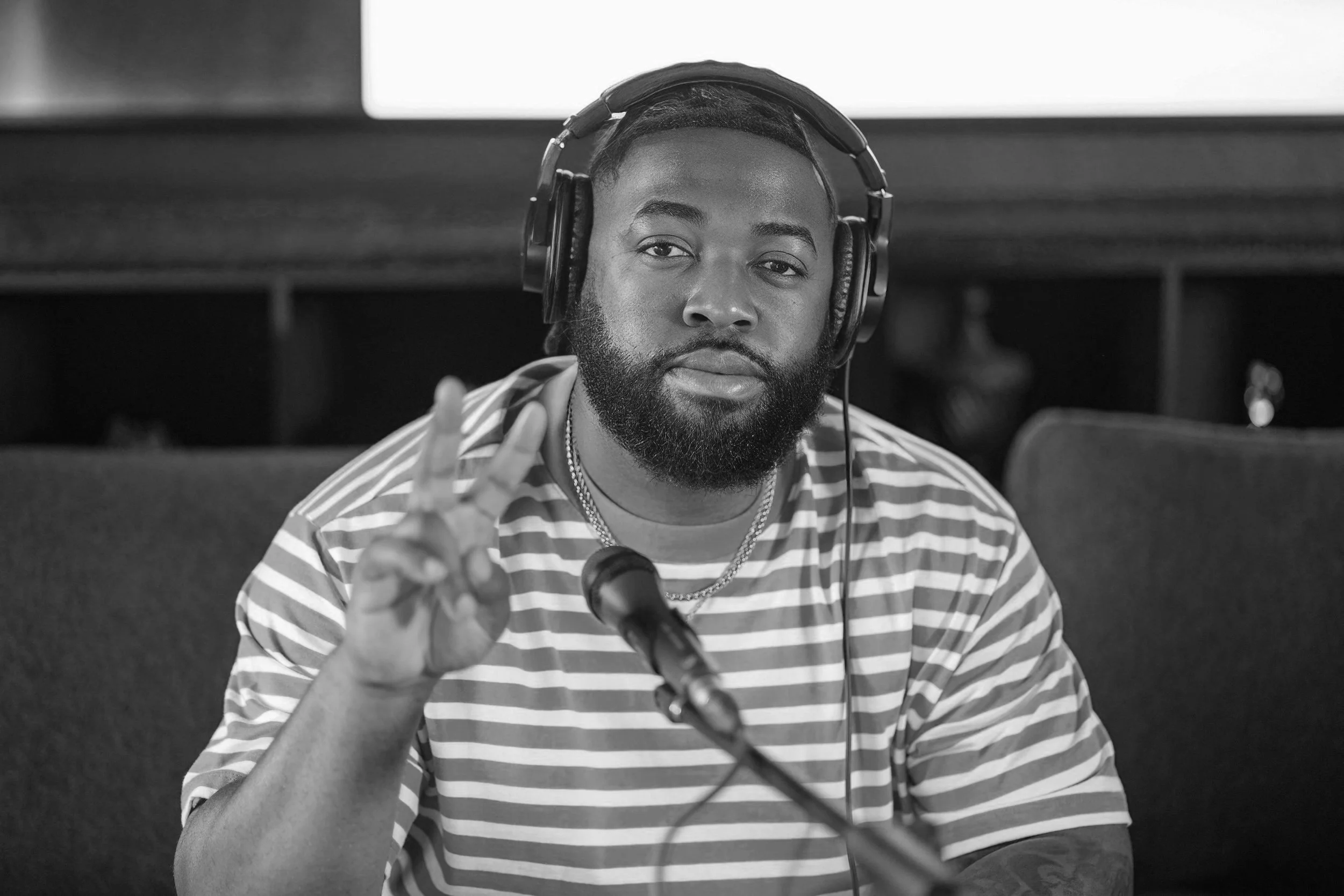 Man in striped shirt wearing headphones and making a peace sign in front of a microphone.