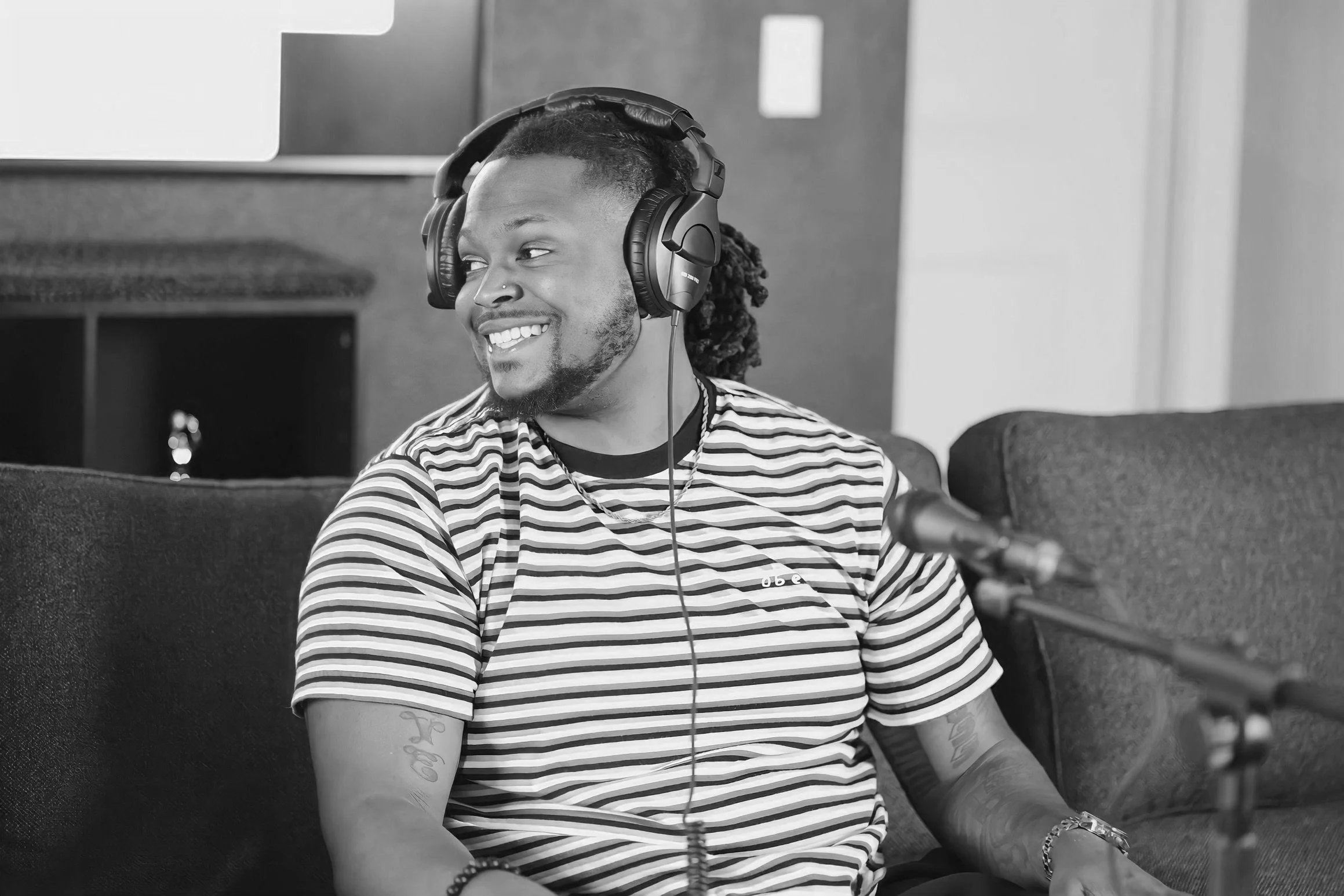 Person with headphones smiling and sitting on a sofa, microphone in foreground, black and white image.