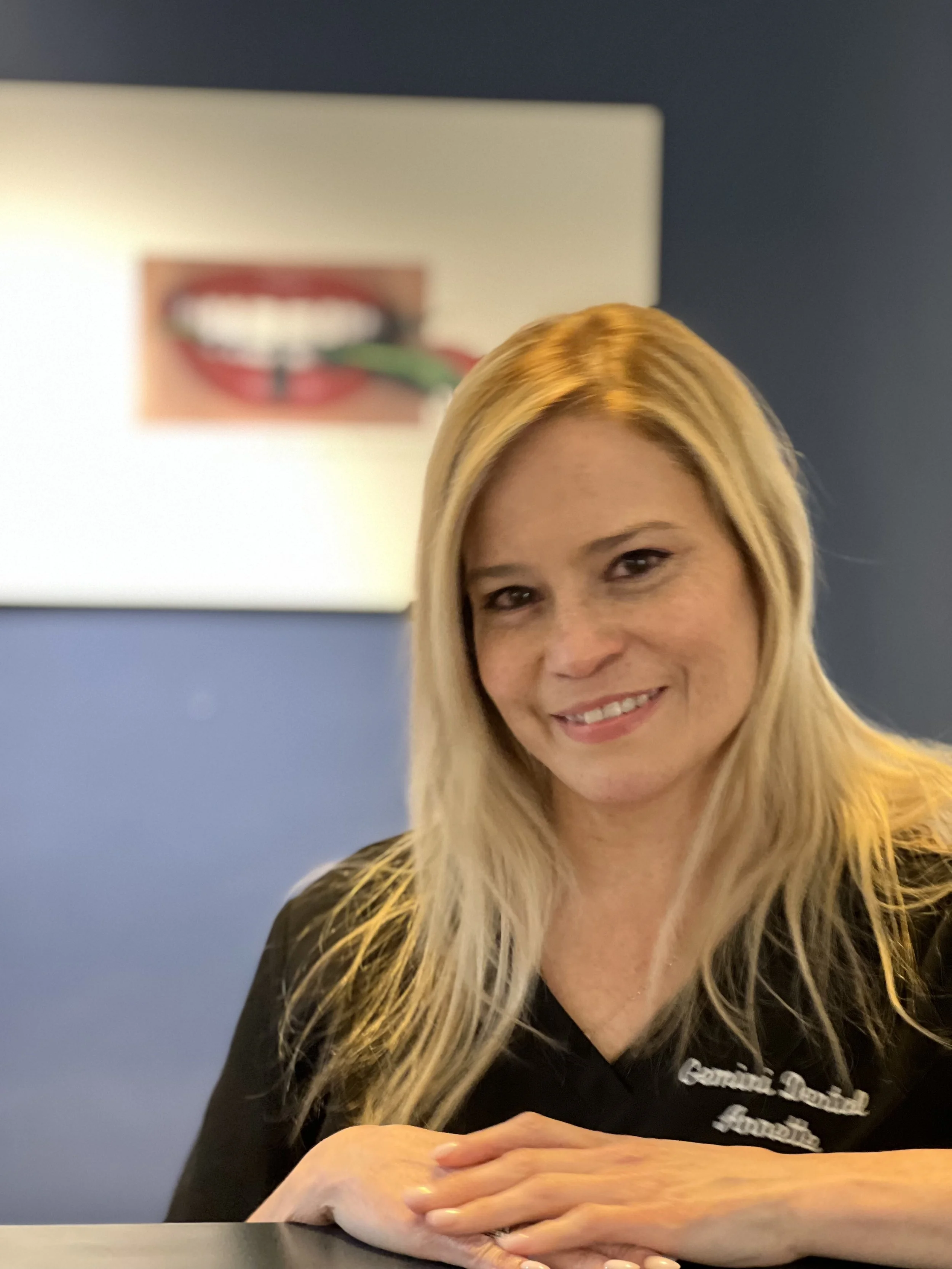 A woman with long blonde hair smiling, sitting with her hands folded on a table, in front of a wall with a blurred poster of a mouth with teeth and a green tongue.