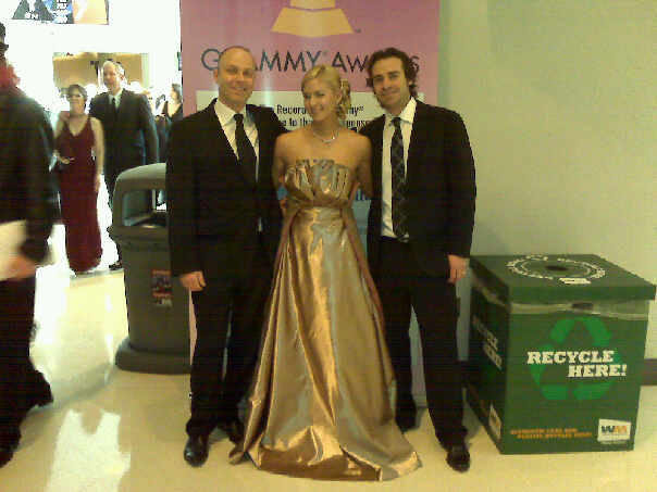 Shannon Smadella at the Grammy Awards dressed in formal attire standing in front of a recycling bin and a pink sign at an event, with other guests in the background.