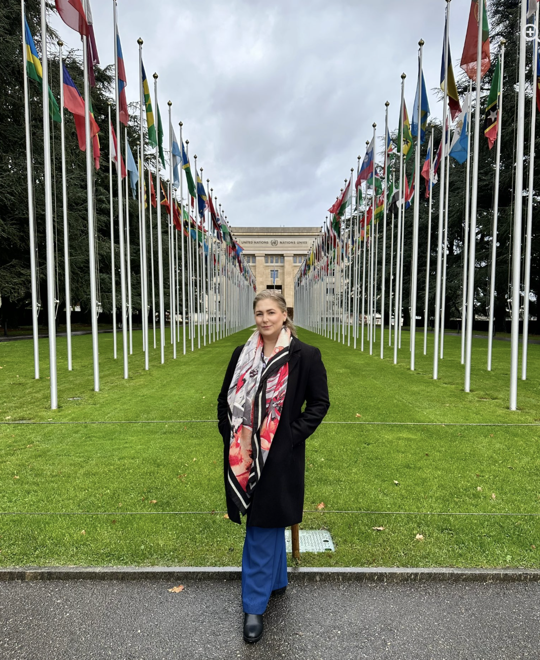 A woman standing in front of the United Nations headquarters in Geneva, Switzerland, with multiple international flags lining the pathway behind her. Shannon Smadella