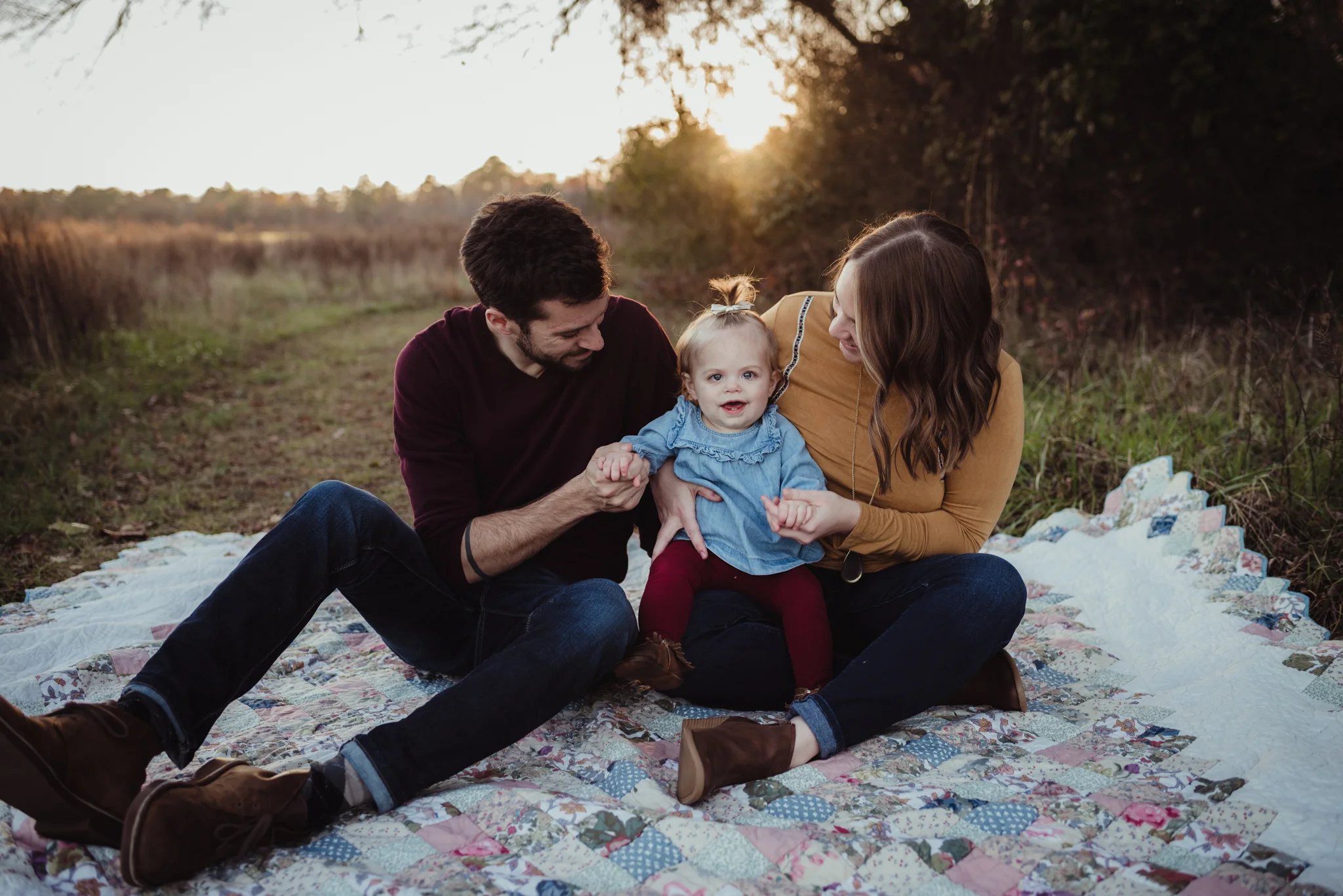 This young family of three sits on an old quilt with their young daughter at sunset during their family photo session with Rose Trail Images at Horseshoe Nature Park in Wake Forest, NC.