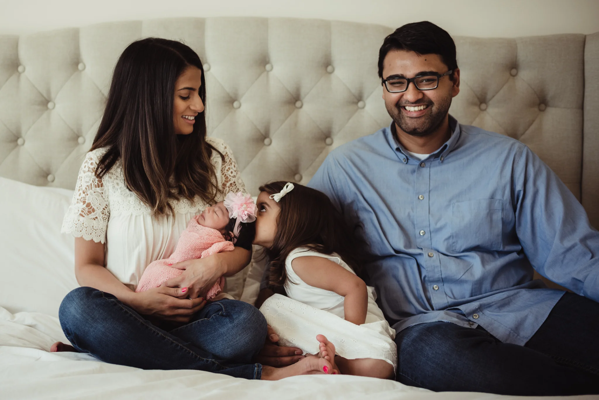 The new family of four sit on the bed while older sister kisses baby sister during their lifestyle newborn session with Rose Trail Images in Raleigh, North Carolina. 