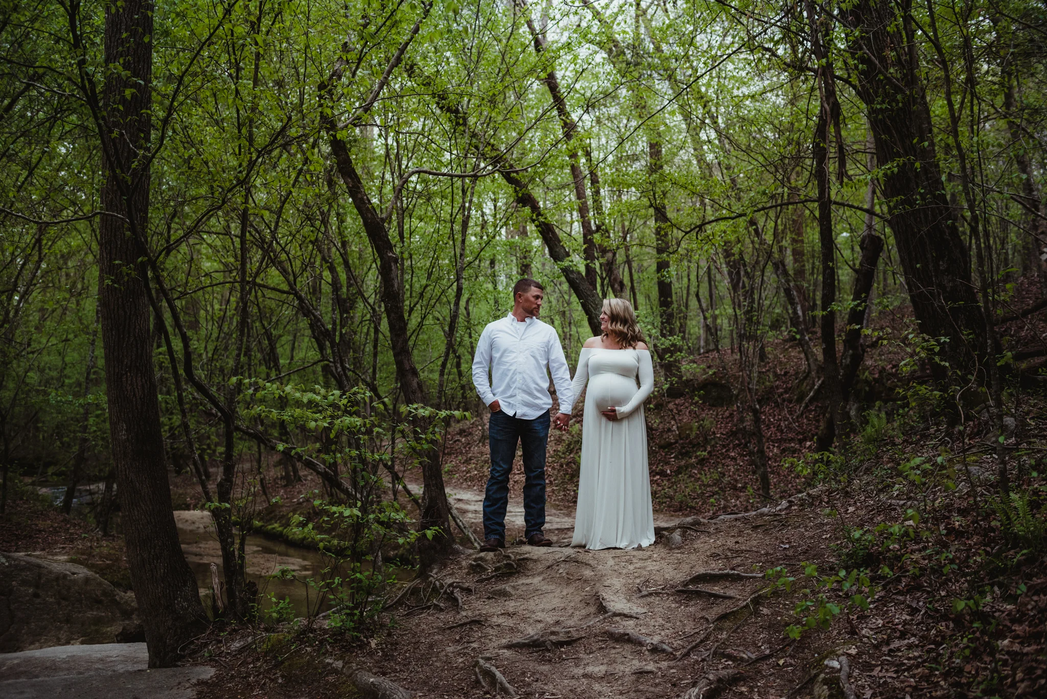 The soon to be parents stand side by side on the wooded trail during their maternity shoot with Rose Trail Images at Mill Bridge Nature Park in Rolesville, NC.