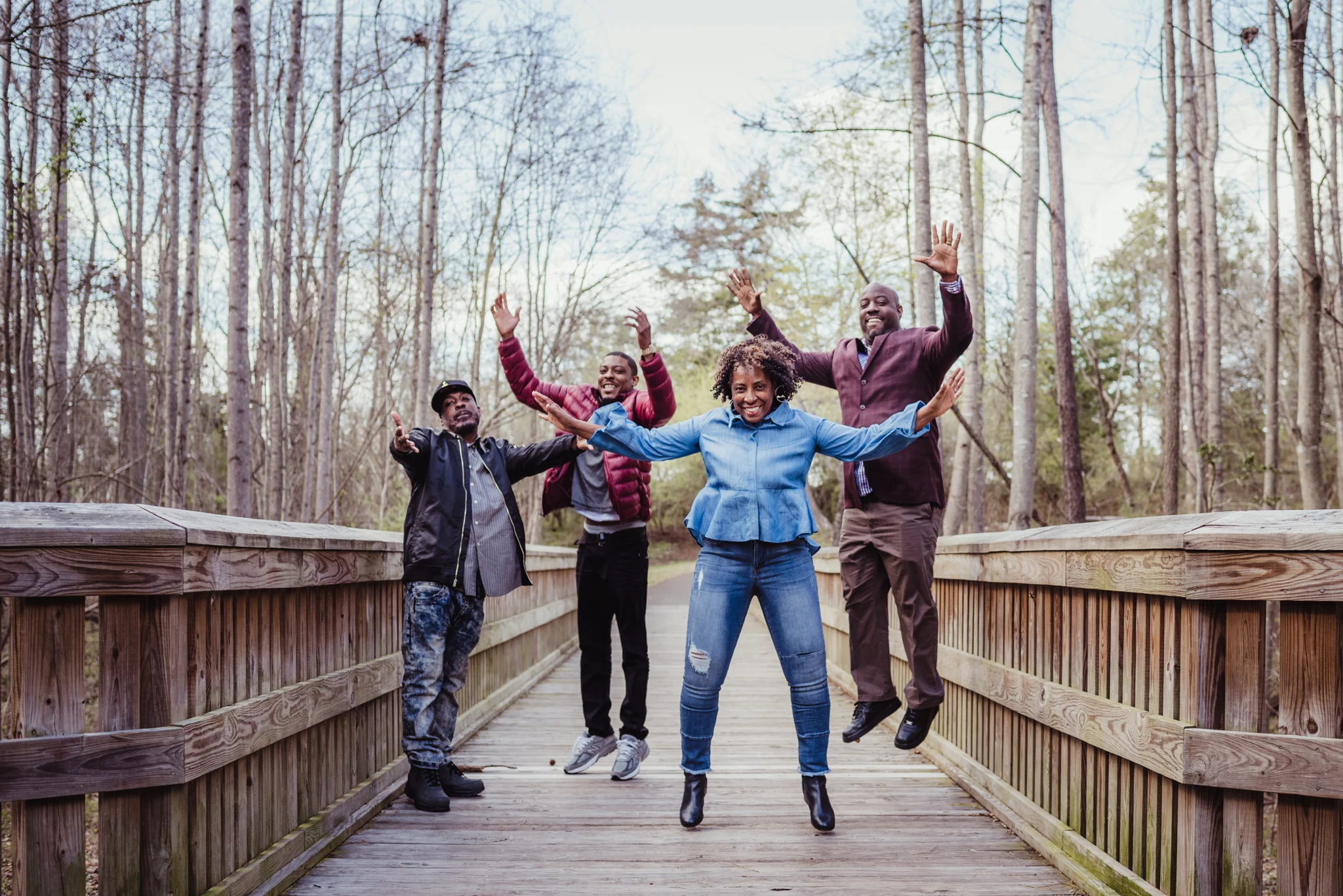 Mom and her adult sons jump for joy on the wooden bridge in Rolesville Main Street Park during their family photo session with Rose Trail Images.