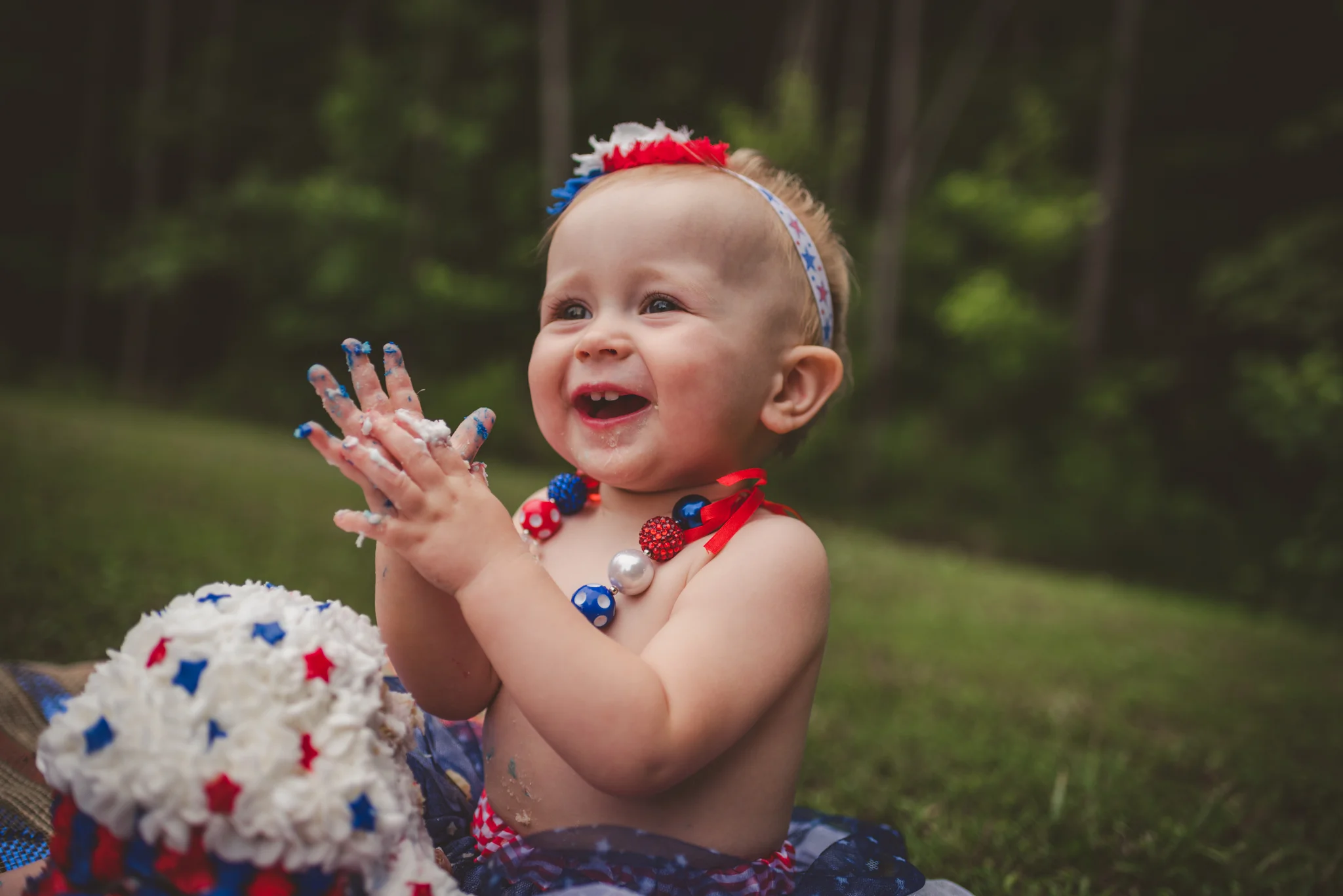This little girl claps while eating red, white, and blue cake for her one year old fourth of July cake smash session with Rose Trail Images in Rolesville, North Carolina.