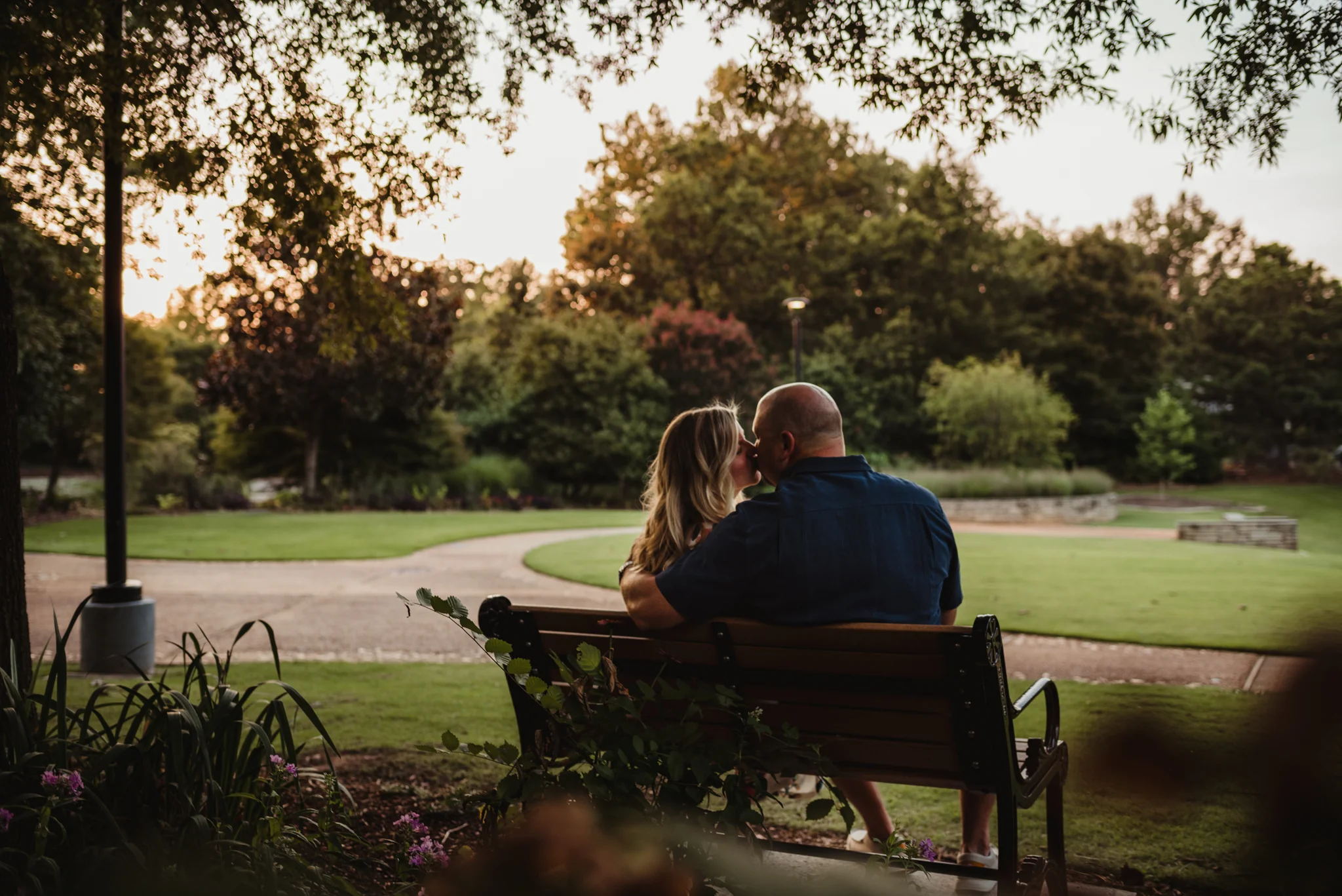 The bride and groom sneak a kiss on the bench during their engagement session at Fred Fletcher Park in Raleigh, North Carolina with Rose Trail Images.
