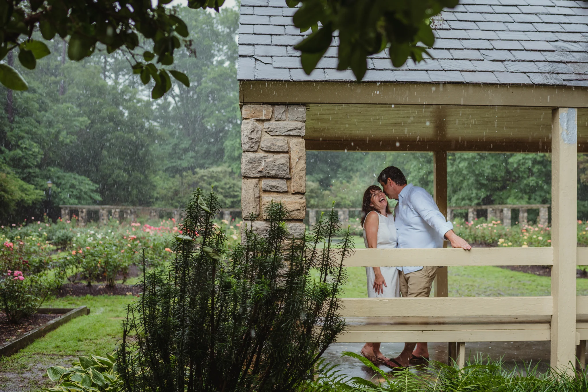 The bride and groom to be laugh together under the shelter during the rainstorm in Raleigh, during their engagement session with Rose Trail Images.