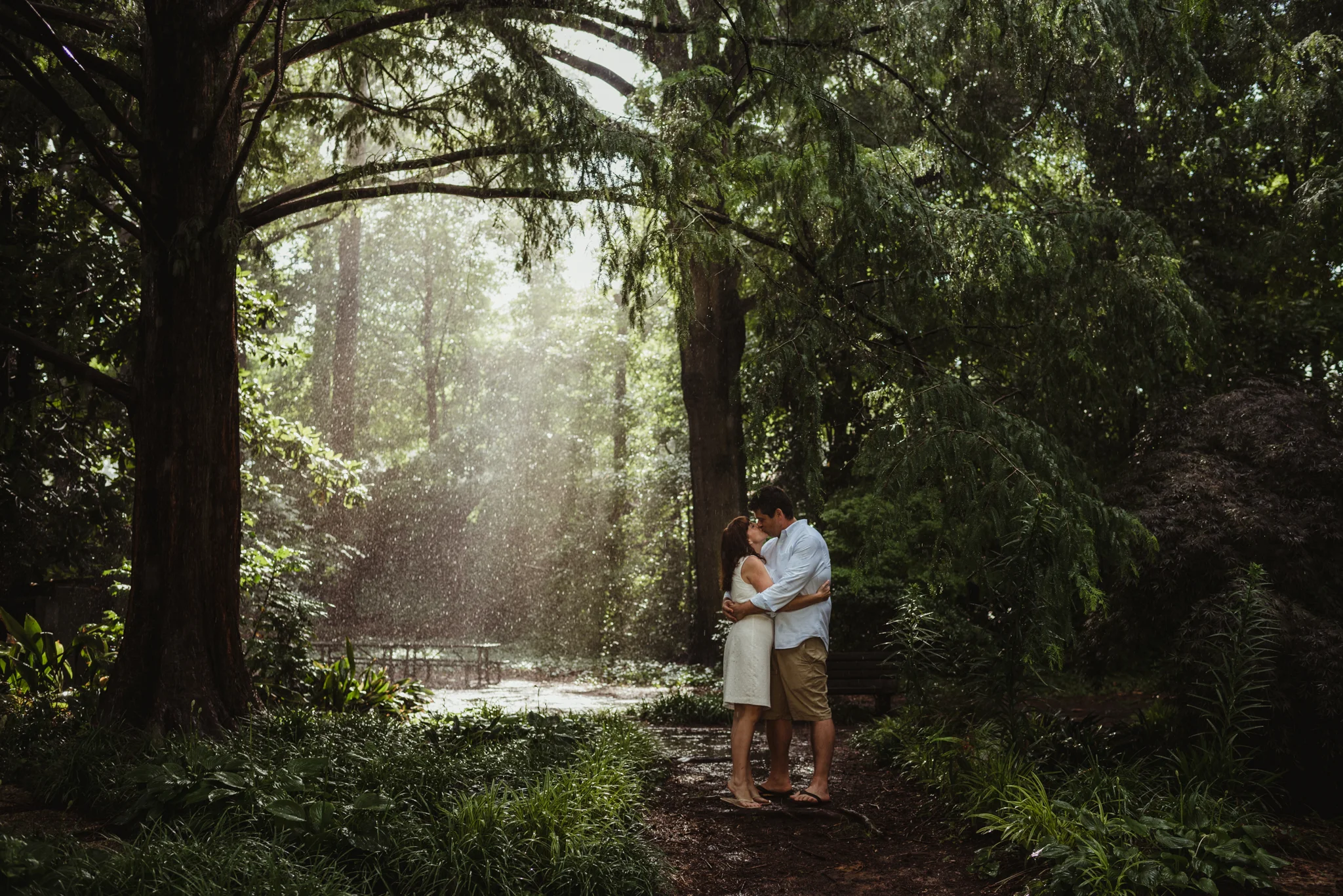 The bride and groom to be kiss in the rain and sunray during the rainstorm in Raleigh, during their engagement session with Rose Trail Images.