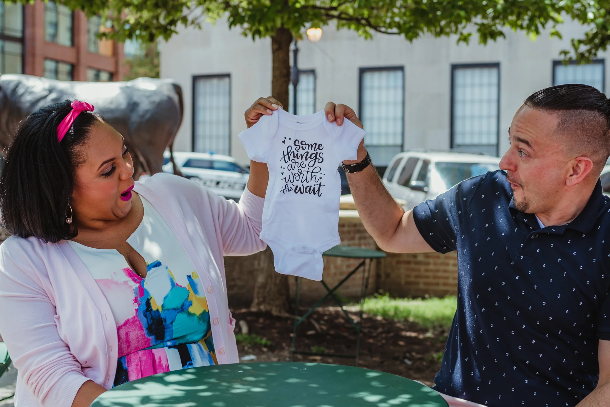 The parents to be hold a onesie saying “some things are worth the wait” outside in Durham during their baby announcement photo session with Rose Trail Images.
